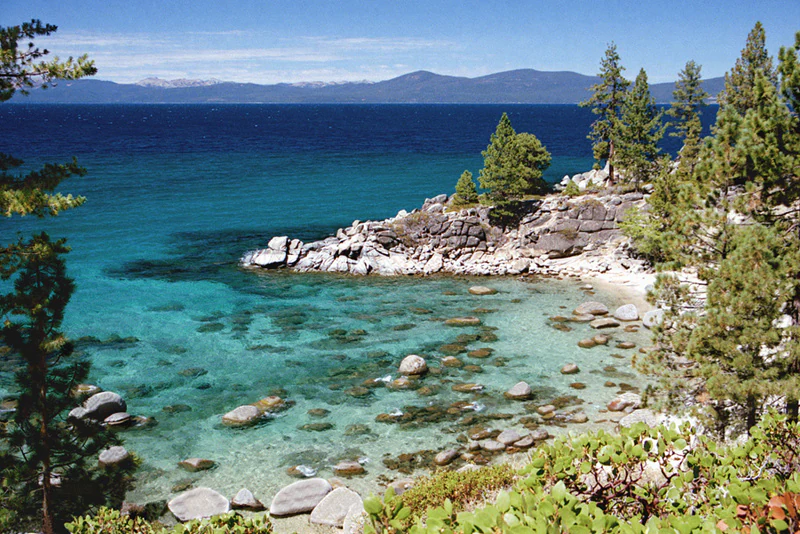 A high-angle view of a small, rocky cove with clear, turquoise water, surrounded by pine trees and large boulders, with a large, deep blue lake and distant mountains in the background.