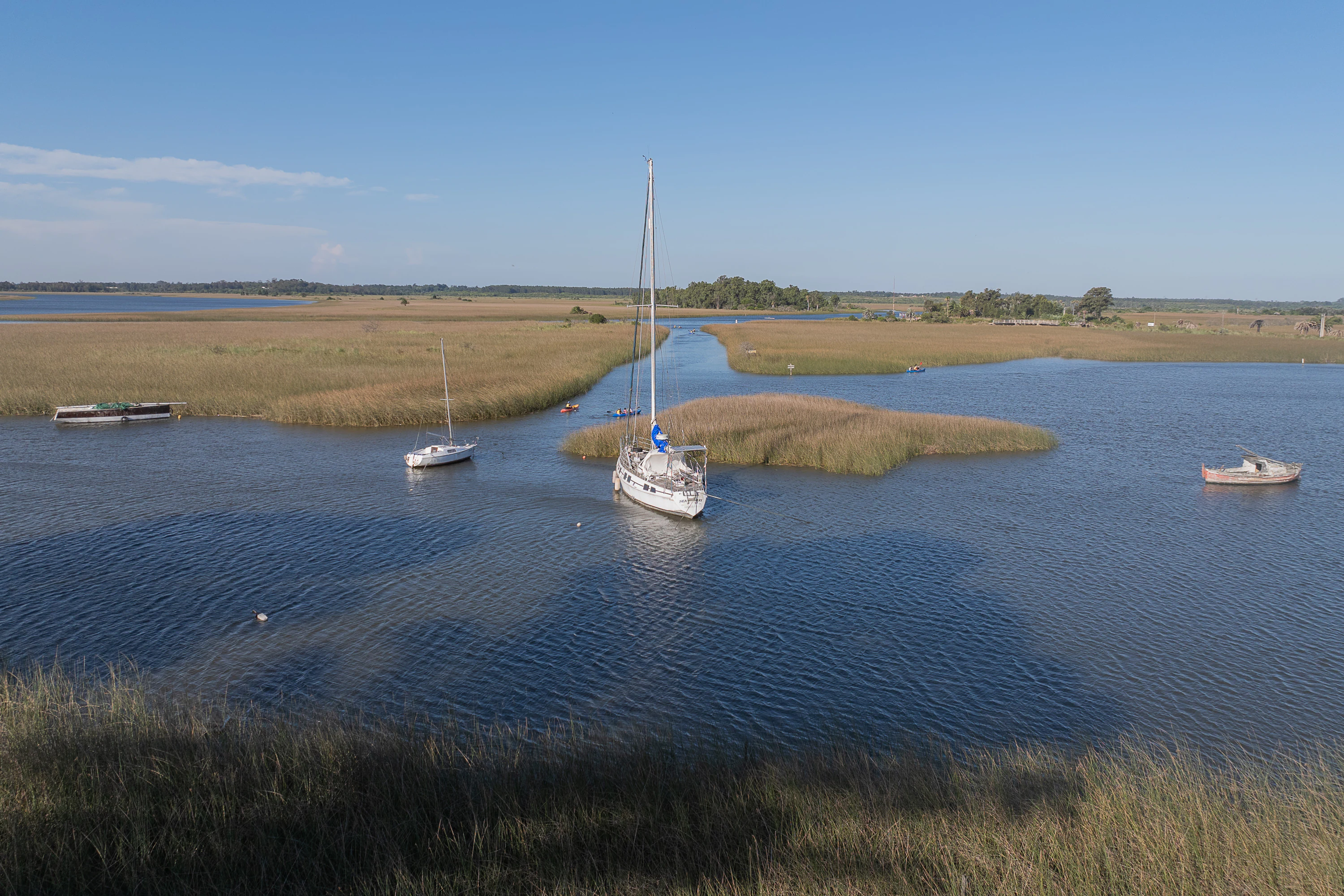 An aerial drone view of the Santa Lucia Wetlands shows a calm waterway winding through marshland with three small boats and sailboats anchored or floating.