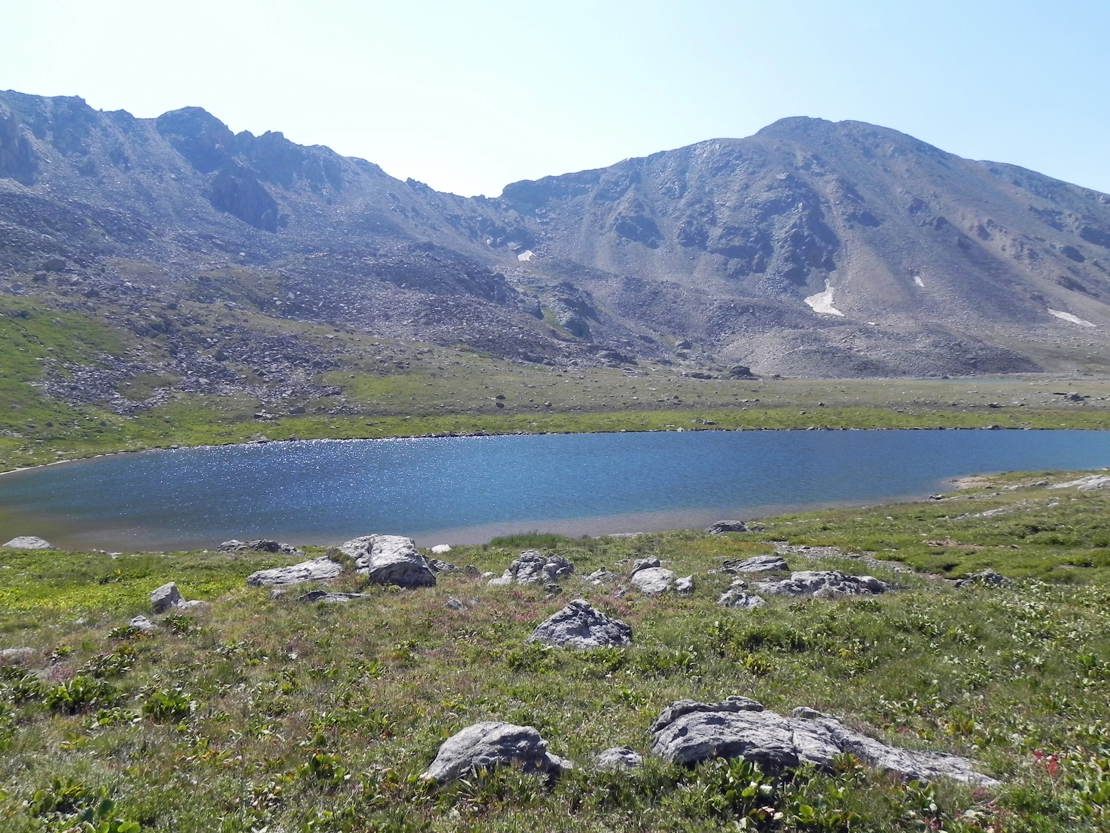 A high-altitude alpine lake sits in the foreground with rocky tundra vegetation and boulders; behind the lake, a large, barren mountain ridge rises into a clear blue sky.