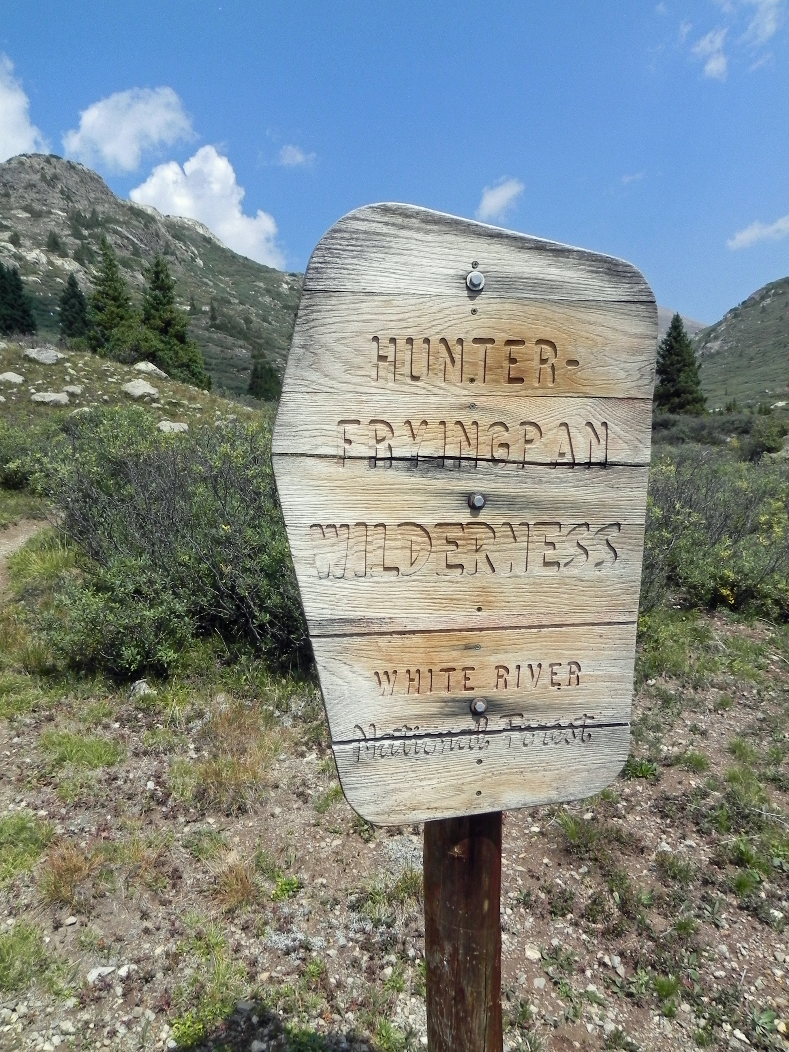 A weathered wooden sign with routed text identifying the Hunter-Fryingpan Wilderness and White River National Forest, set against a mountain landscape with green slopes.