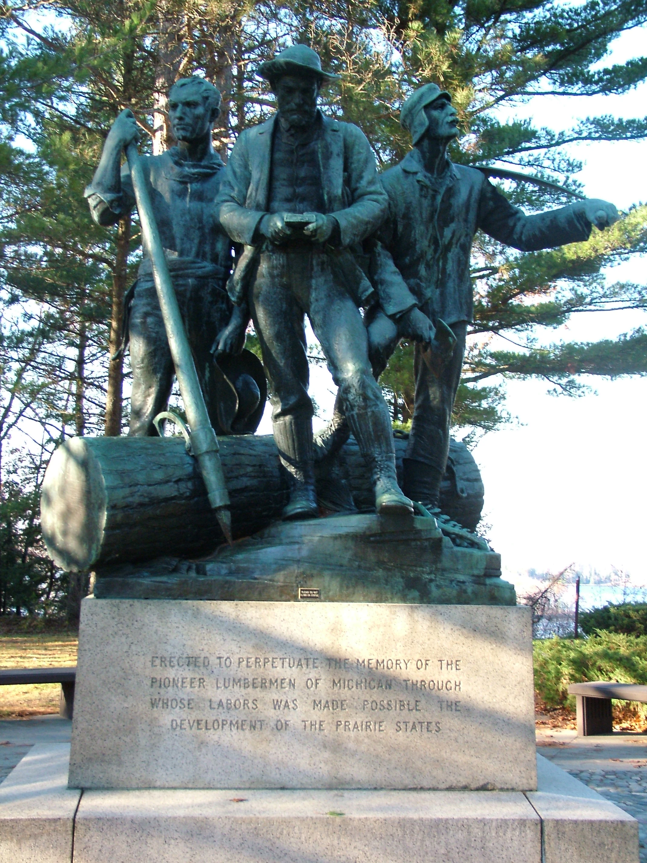 A bronze statue featuring three lumberjacks standing around a log, set on a stone pedestal with an engraved inscription, surrounded by pine trees.