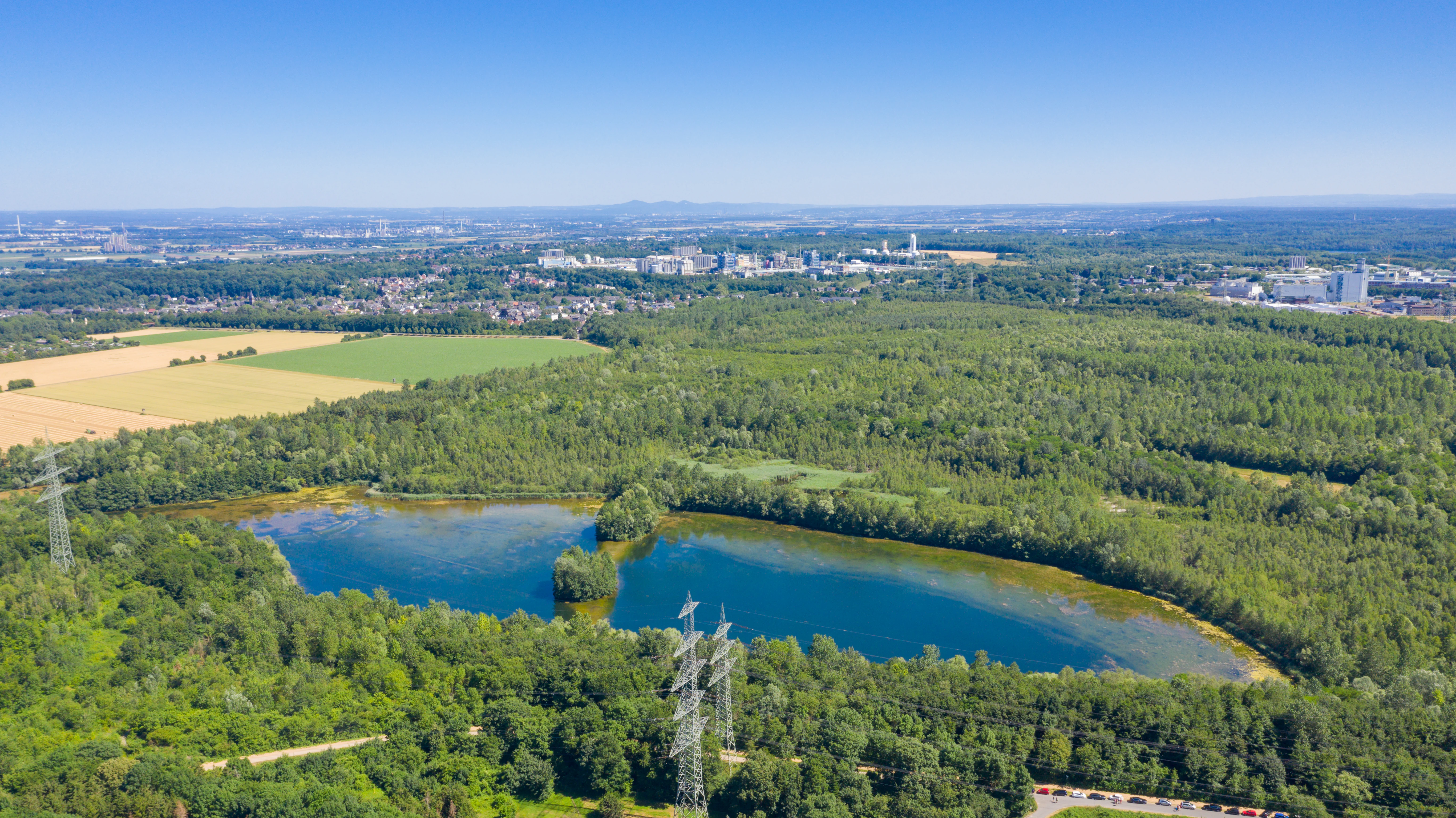 An aerial drone view showing a small blue lake nested within a dense green forest, with power line towers in the foreground and urban industrial buildings in the background.