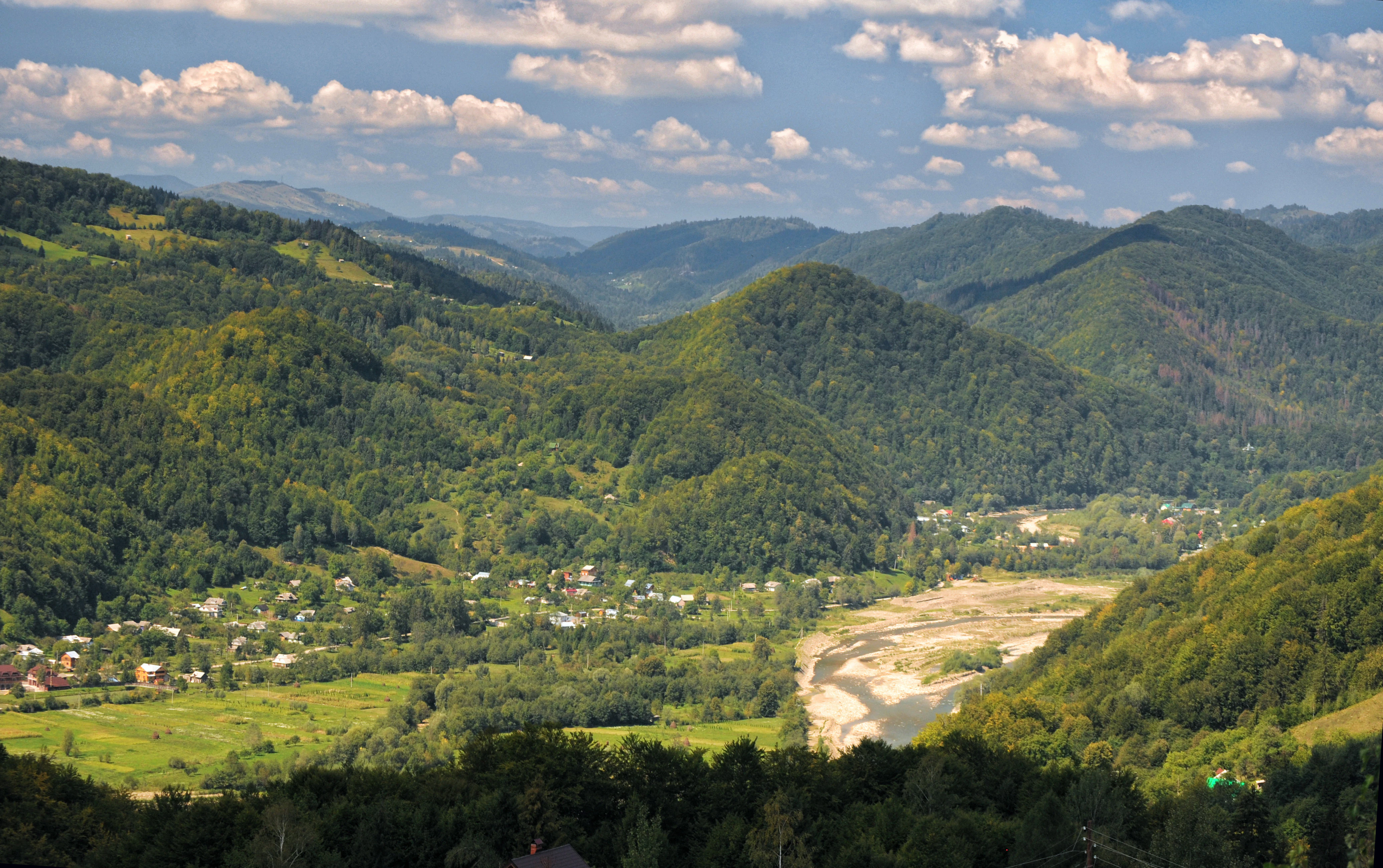 Mountainous landscape with green valleys, a river, and distant hills under a partly cloudy sky