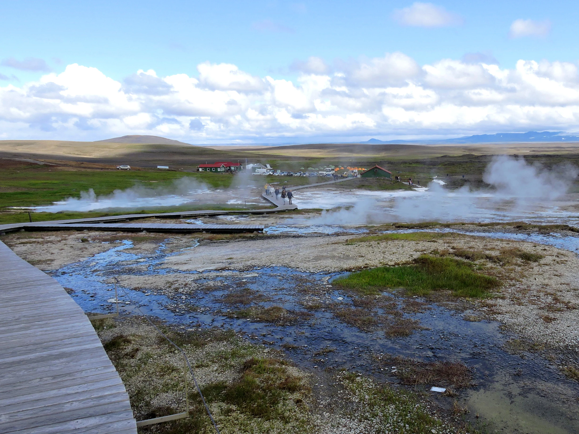 A wide view of the Hveravellir geothermal area in Iceland, featuring a wooden boardwalk path stretching across a marshy, steaming landscape with low rolling hills in the background.