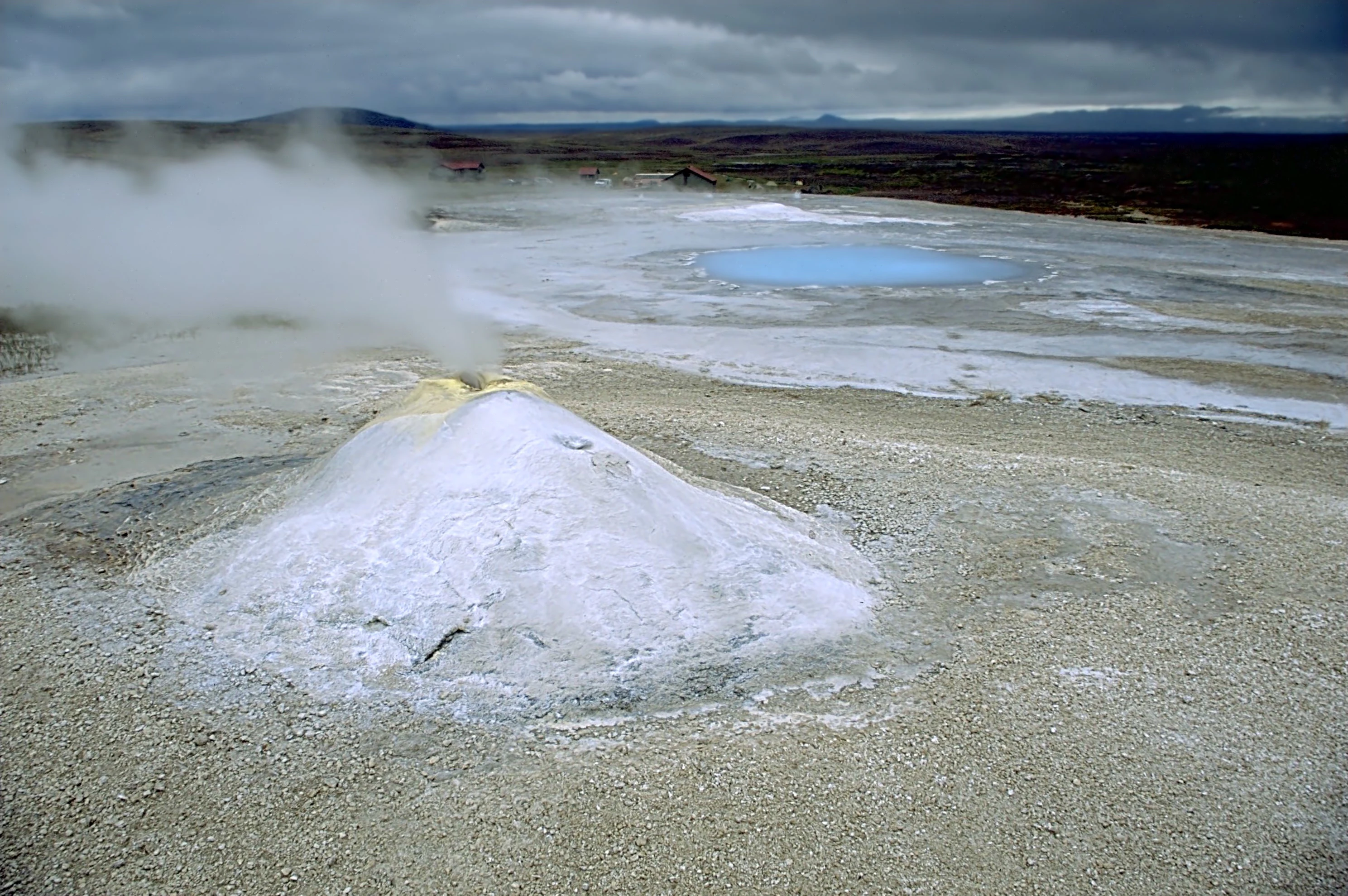 A wide angle view of a white, conical geothermal vent emitting steam, next to a small, bright blue hot spring pool on a flat, barren landscape under a cloudy sky.