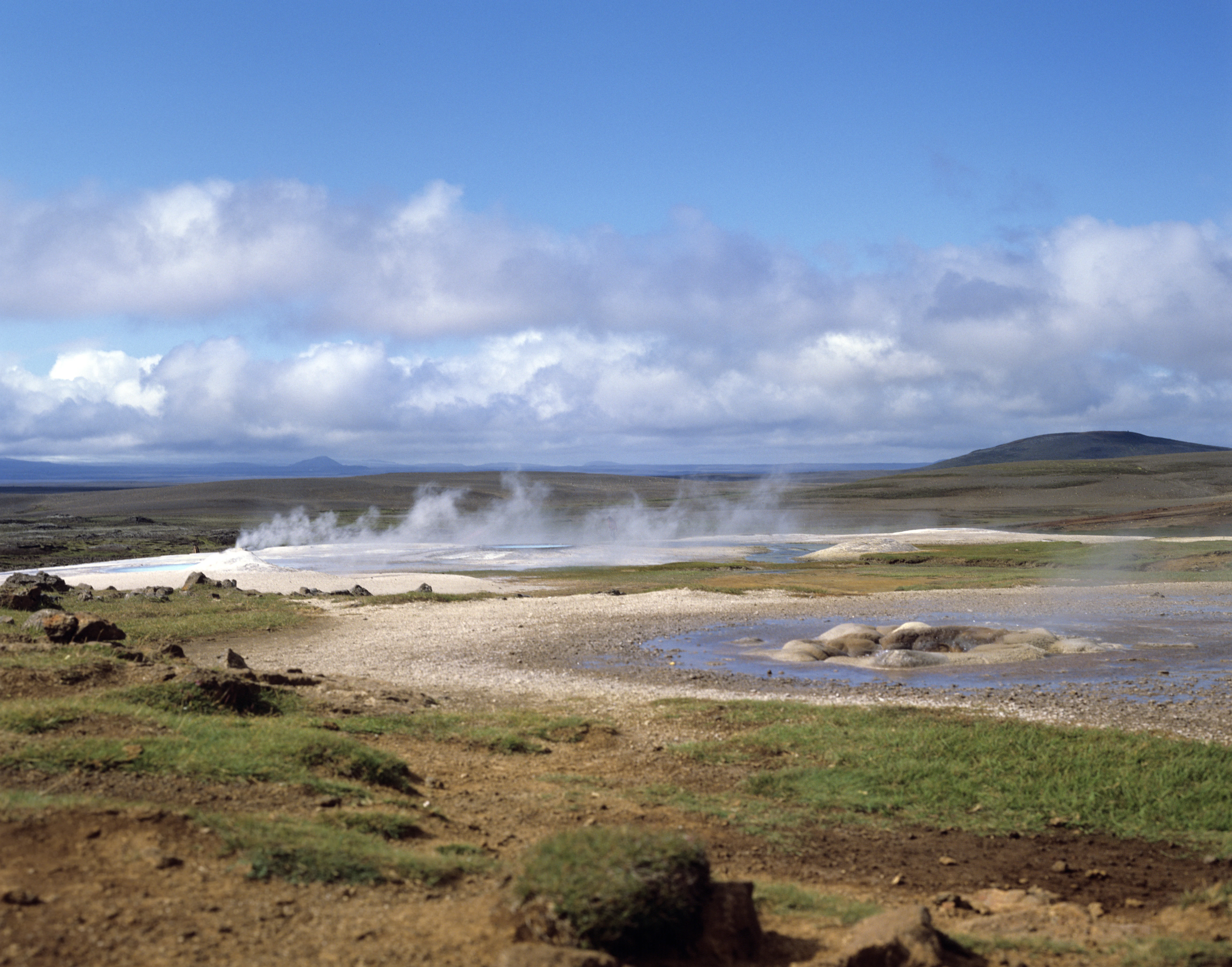 A wide view of the Hveravellir geothermal area in Iceland, with steam rising from vents and hot springs across a flat, sparse, and rocky landscape under a blue sky with scattered clouds.