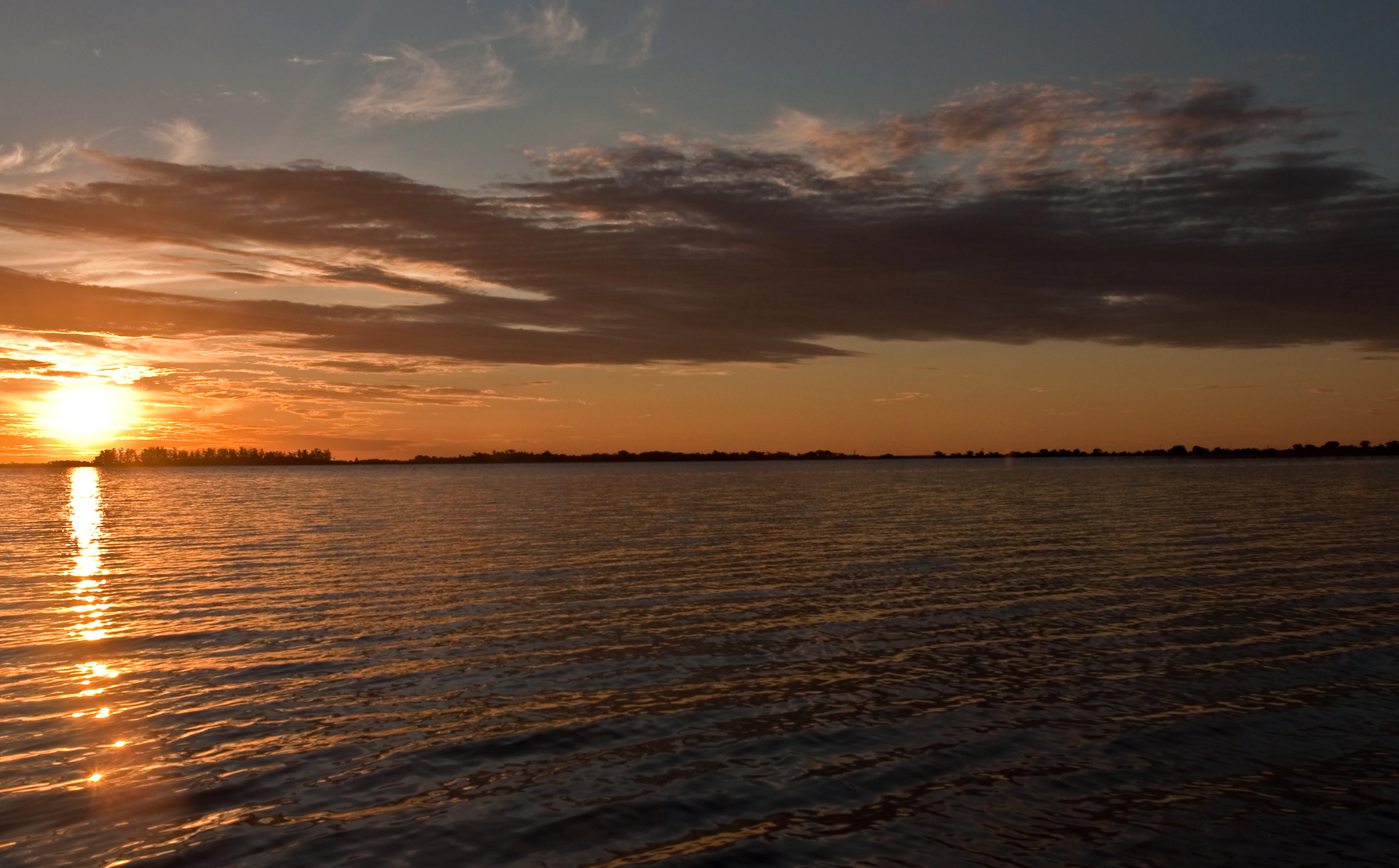 A wide sunset view over calm open water with silhouetted distant shorelines and clouds catching the golden light.