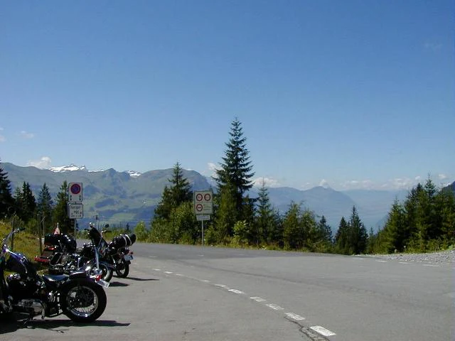 A group of motorcycles parked on the side of a paved road at a mountain pass, with evergreen trees and distant alpine peaks under a clear blue sky.