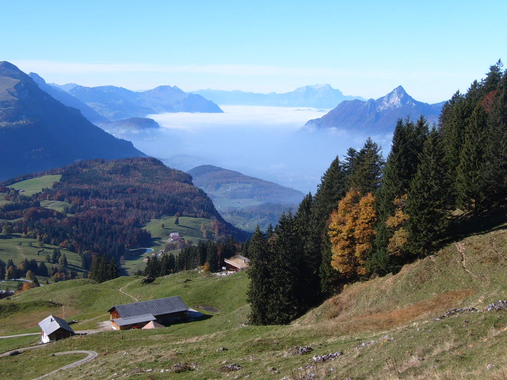 A panoramic alpine landscape showing a layer of fog in the distant valley, surrounded by rolling green hills, coniferous trees, and traditional wooden farm structures in the foreground.