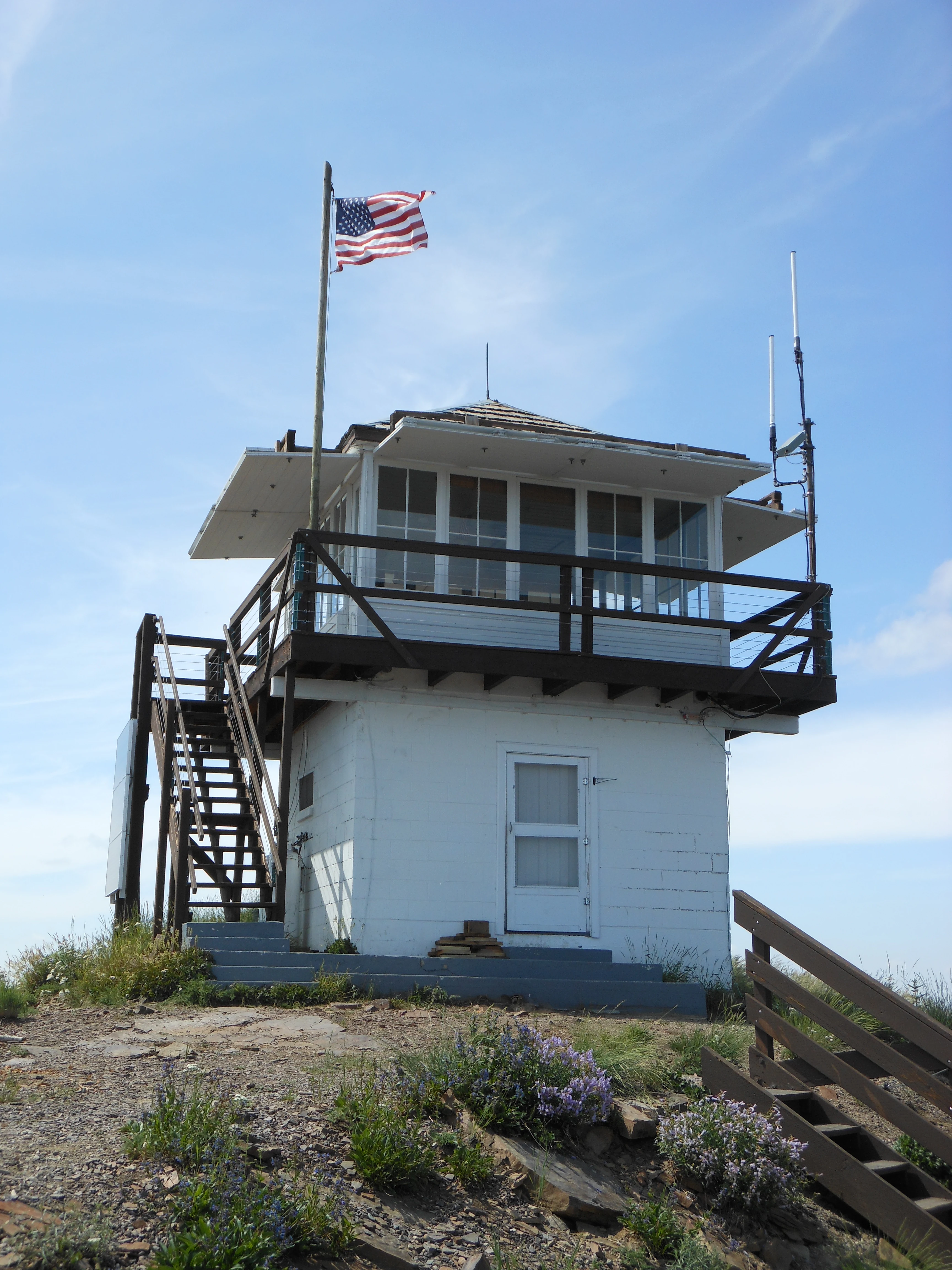 A small, white, square fire lookout tower with a wraparound wooden deck, stairs leading to the entrance, and a flying American flag, set atop a rocky mountain peak against a clear blue sky.