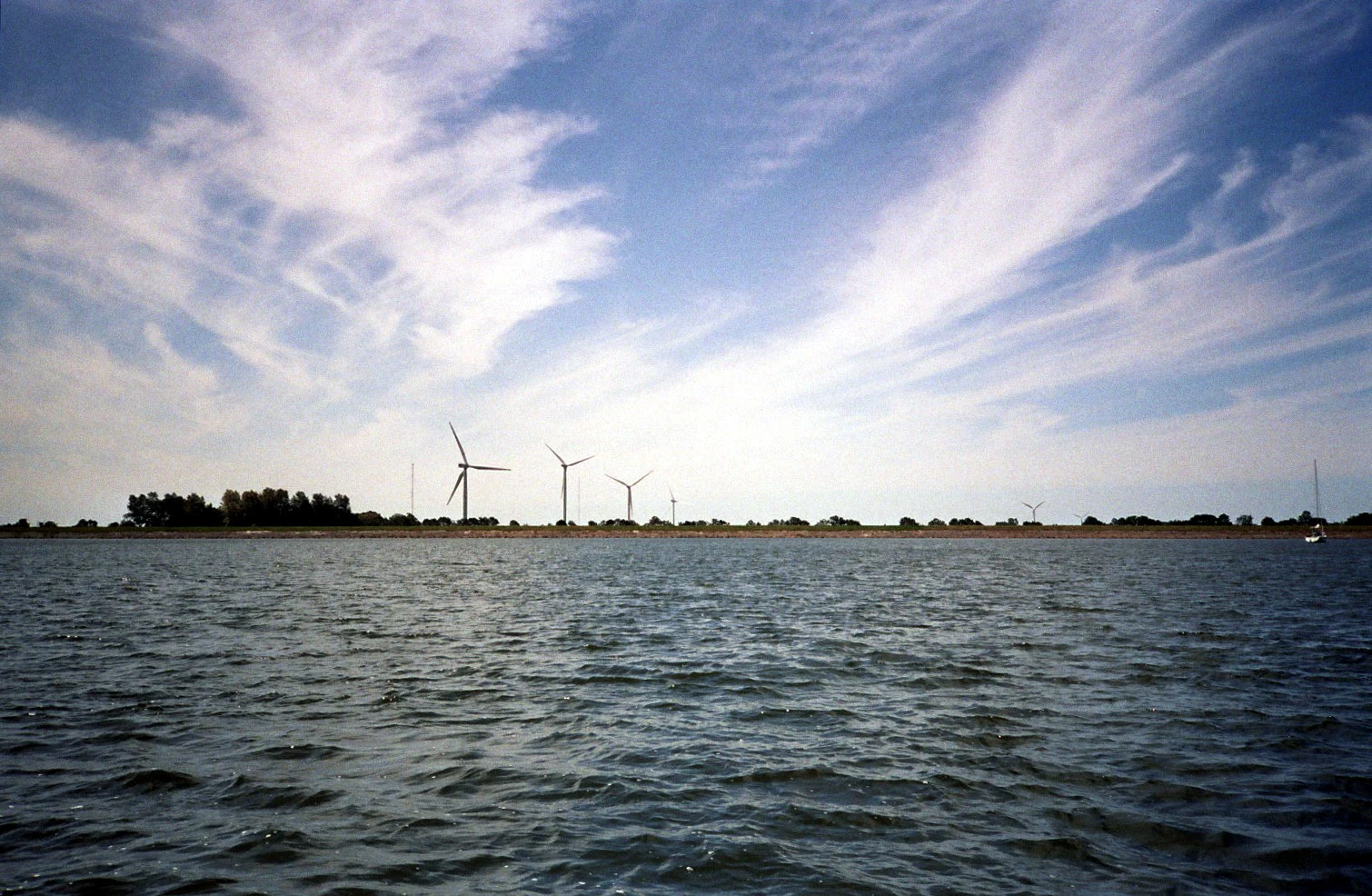 A wide shot of the IJsselmeer lake with a line of wind turbines on the distant shoreline under a bright, wispy-clouded sky.