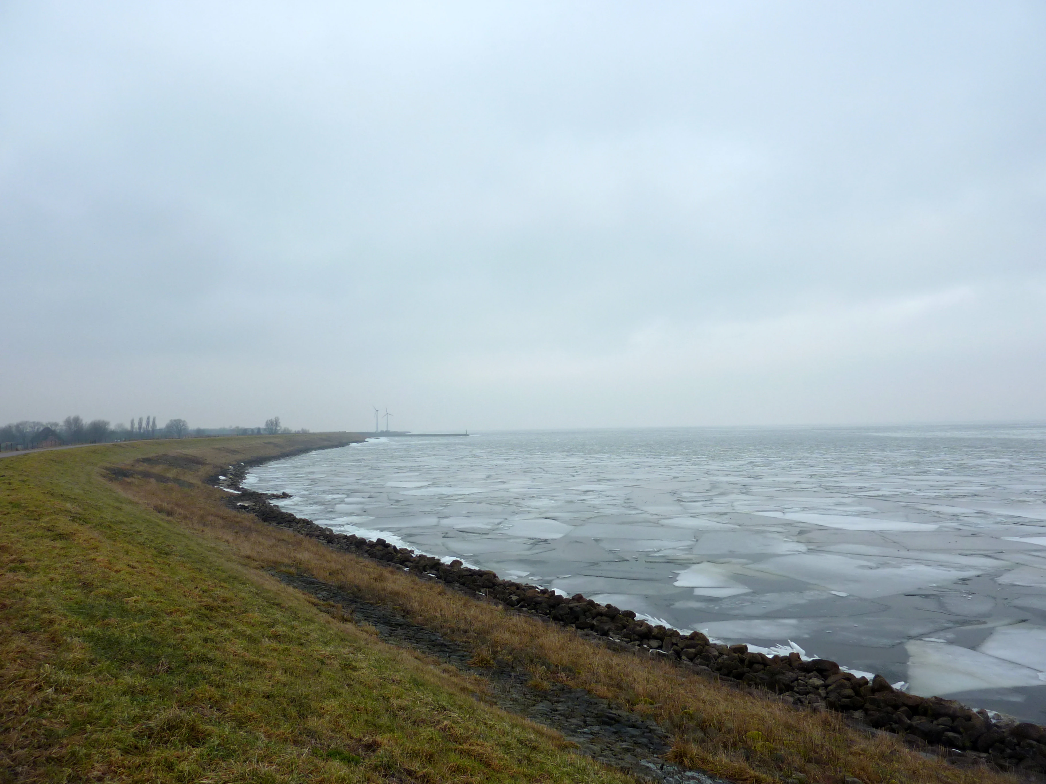 A wide landscape view of a partially frozen lake with ice floes covering the water, viewed from a grassy embankment under a cloudy, overcast sky.