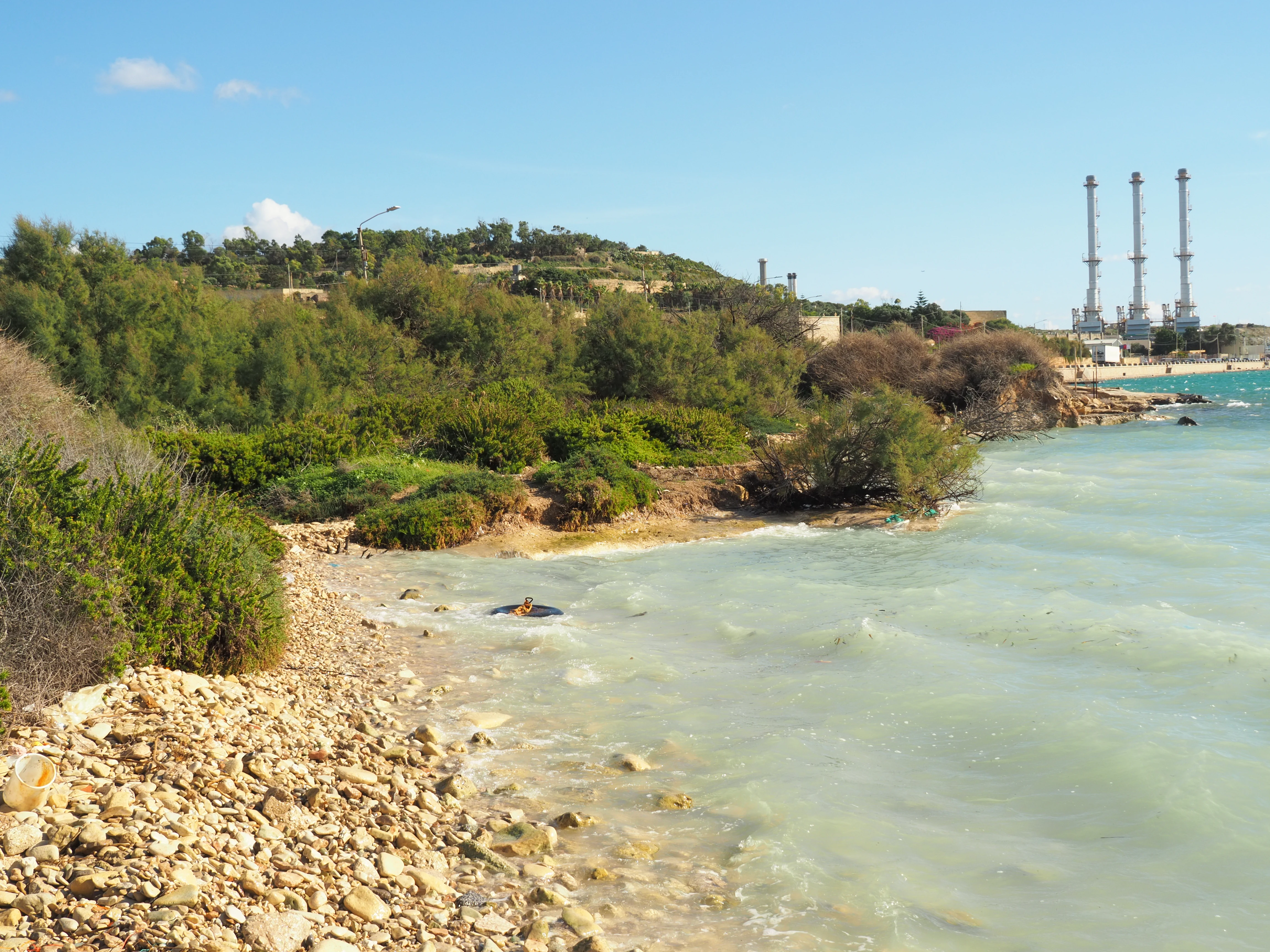 A coastal salt marsh area with rocky shoreline and low vegetation, with the tall stacks of a power plant visible in the distance across the water.