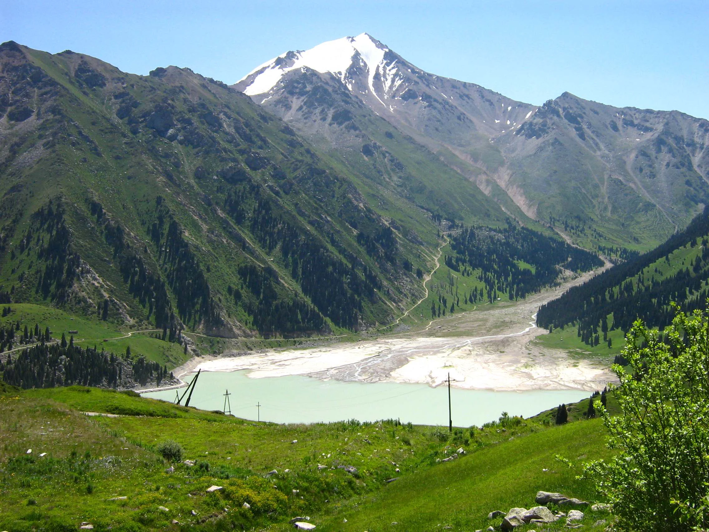Big Almaty Lake with snow-capped mountain peak, green hills, and utility poles in foreground