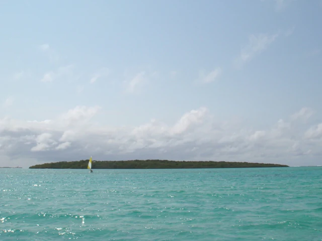 A low-lying tropical island covered in dense green vegetation sits in the middle of a turquoise ocean under a pale sky, with a small sailboat visible near the shore.