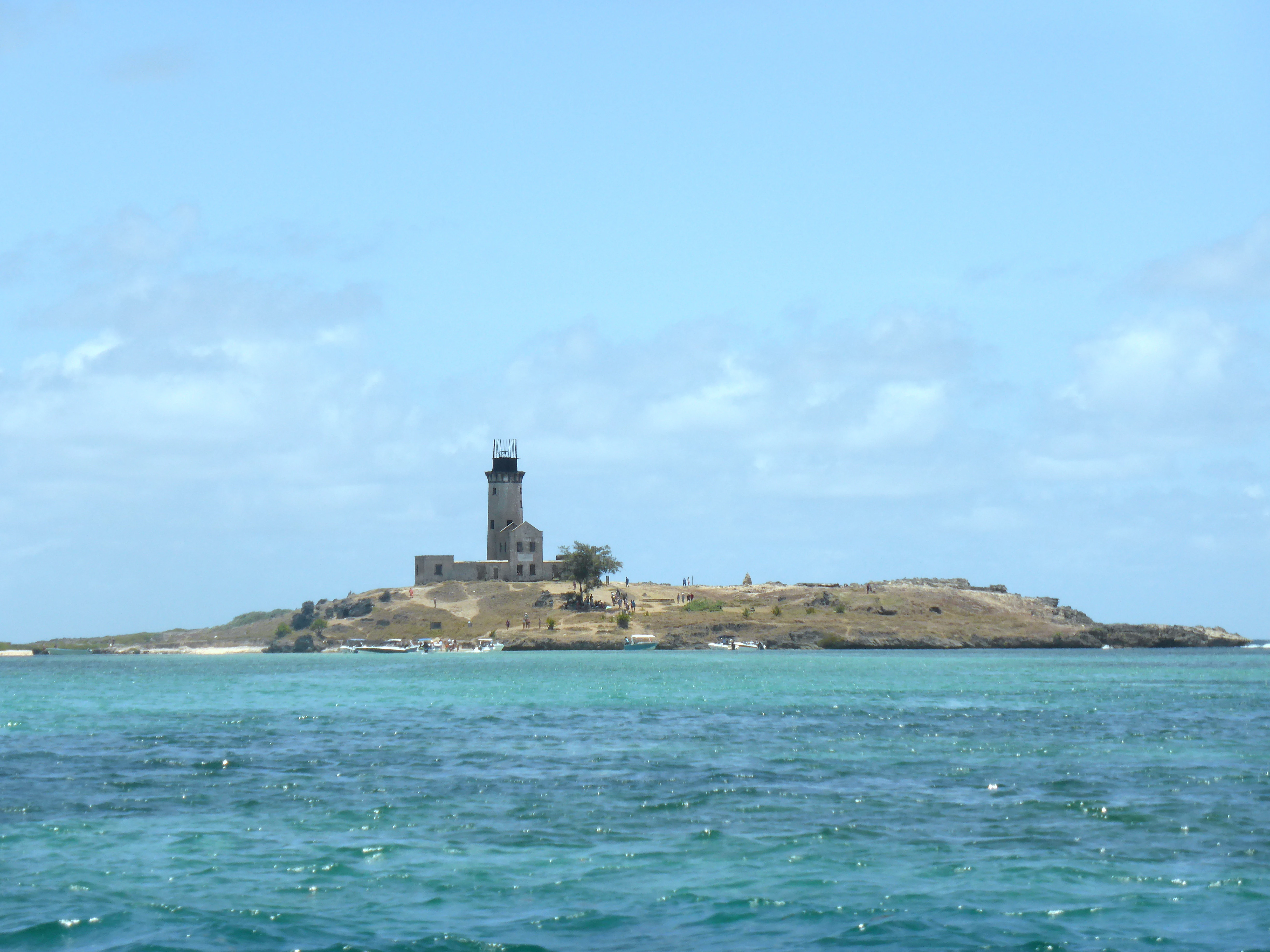 A distant view of Île aux Fouquets island featuring a weathered stone lighthouse, surrounded by turquoise ocean waters under a clear blue sky.