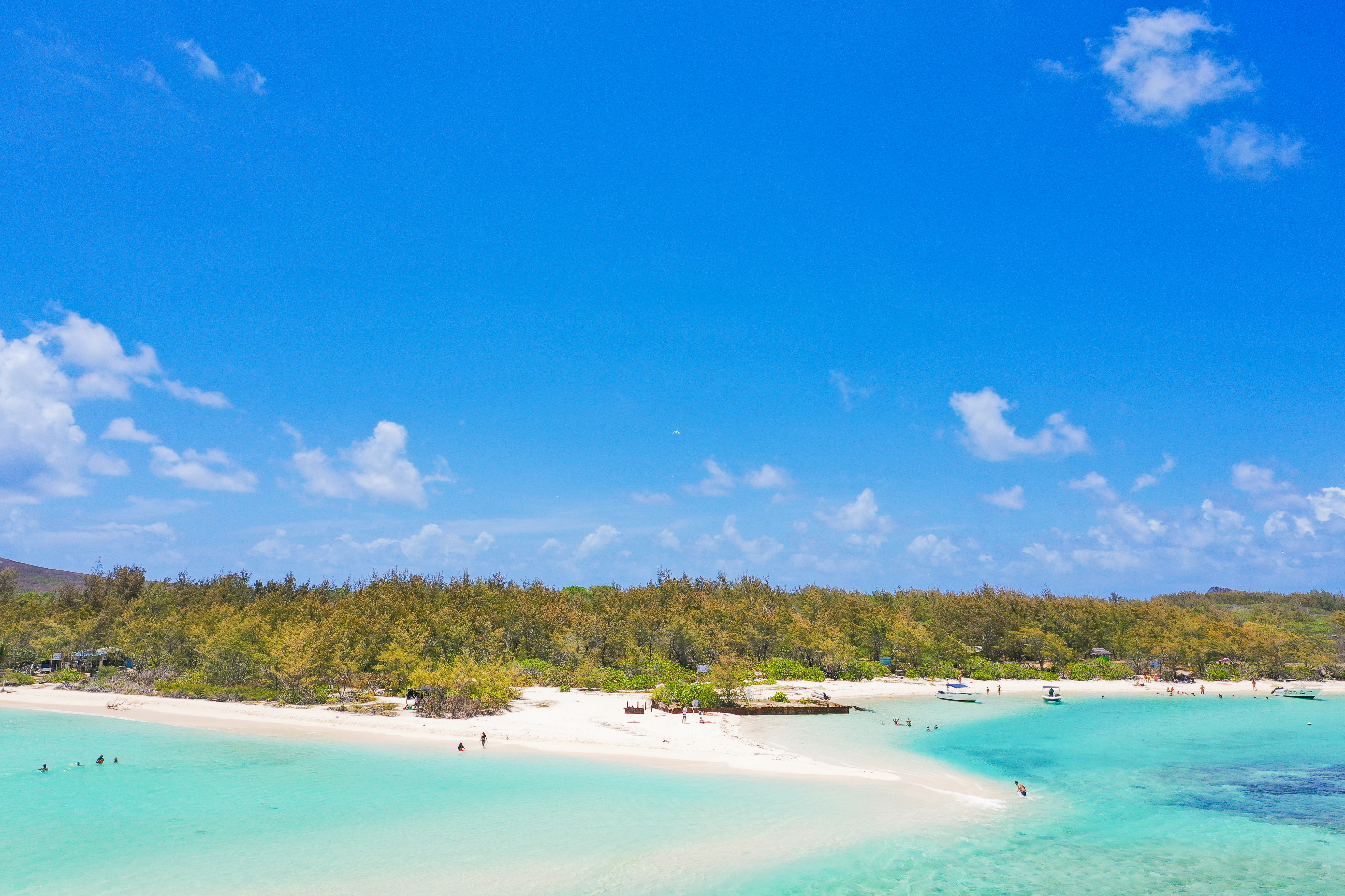A wide aerial view showing a white sandy beach curving into shallow, bright turquoise ocean water on Ile Plate, with low-lying green vegetation and a clear blue sky in the background.
