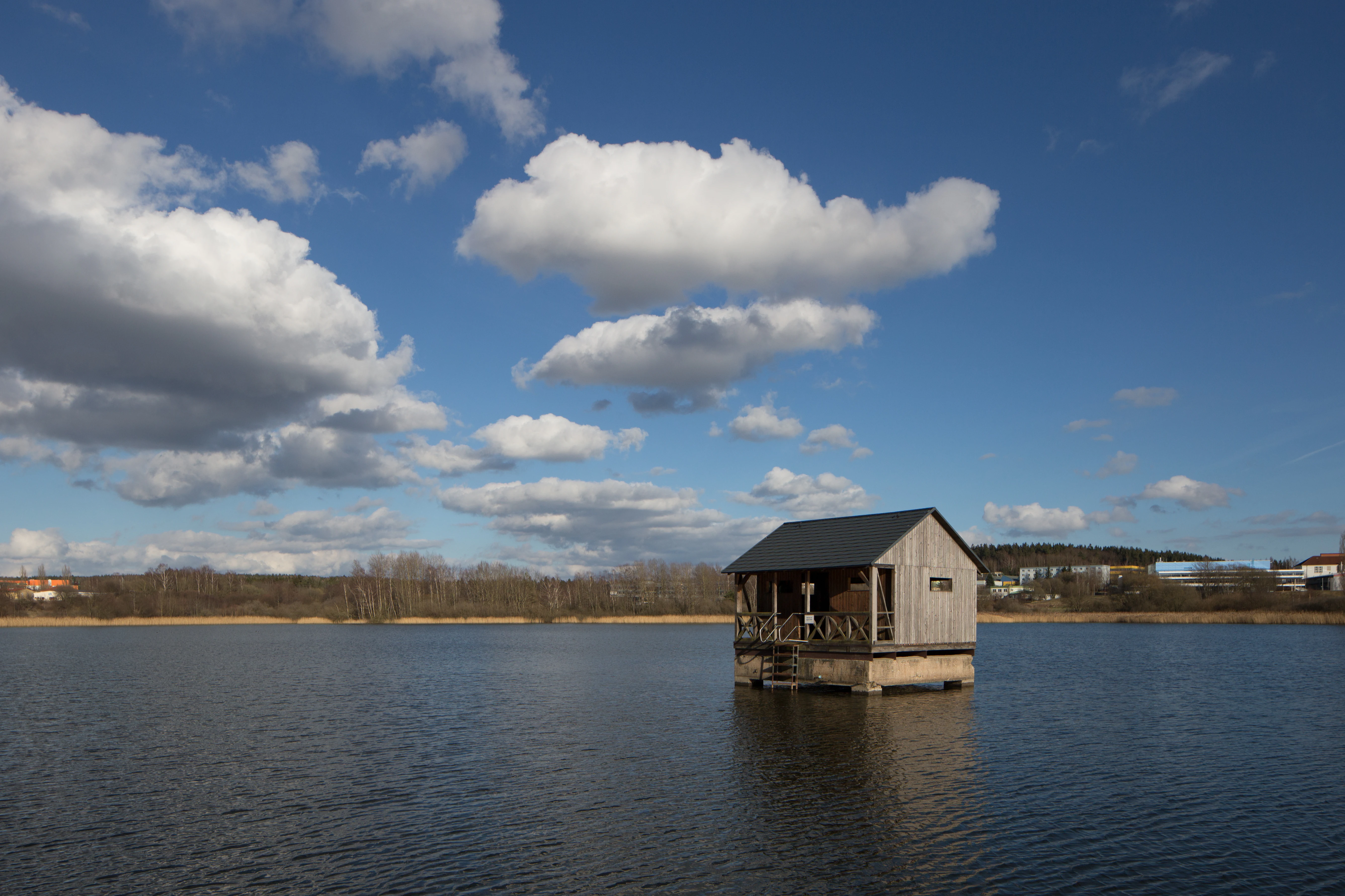 A small wooden stilt house sits in the middle of a calm pond under a bright blue sky with fluffy white clouds, with a distant treeline and buildings on the horizon.