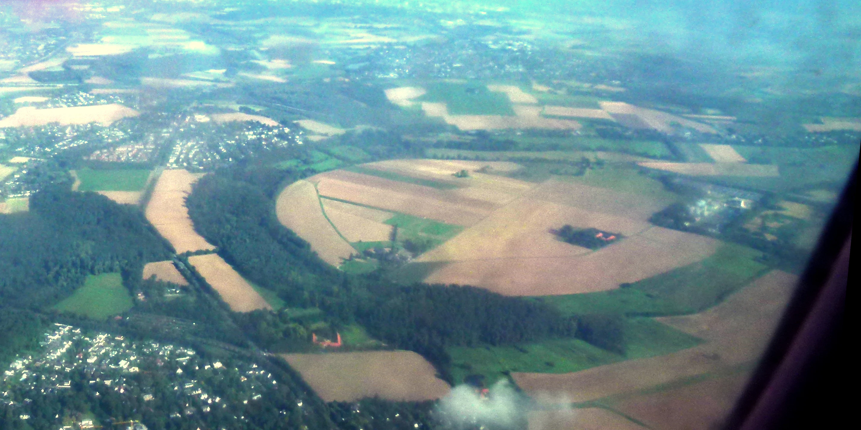 An aerial view from an aircraft window showing the curved, oxbow-shaped forested landscape of the Ilvericher Altrheinschlinge, surrounded by agricultural fields and suburban development.