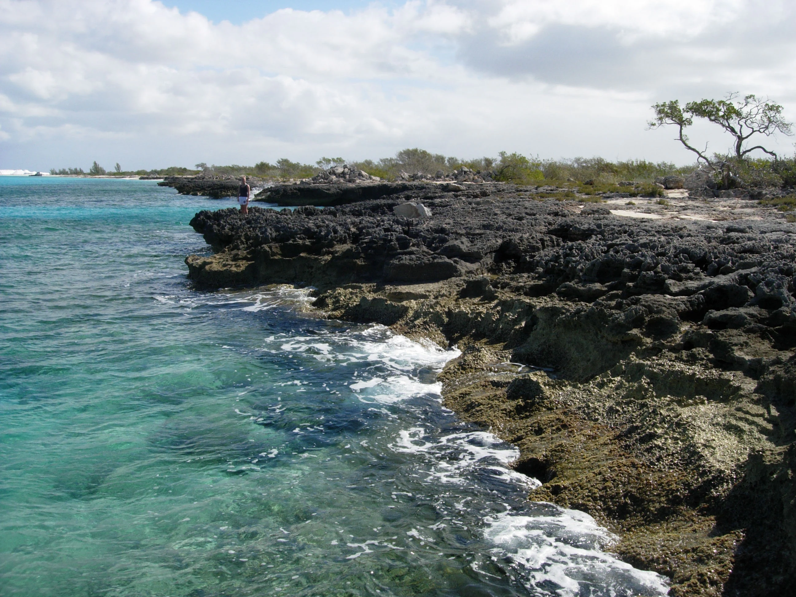 Rocky coastline with turquoise water, white foam, and exposed limestone formations under a partly cloudy sky.