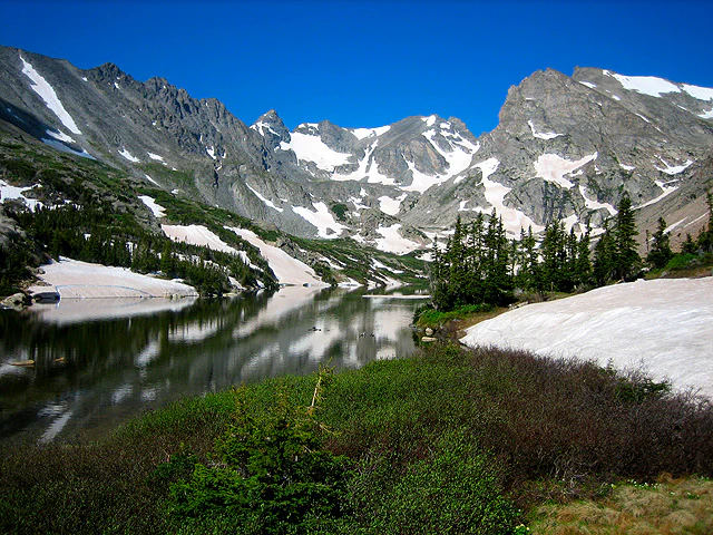 An alpine lake reflecting snow-capped mountain peaks and evergreen trees under a bright blue sky.