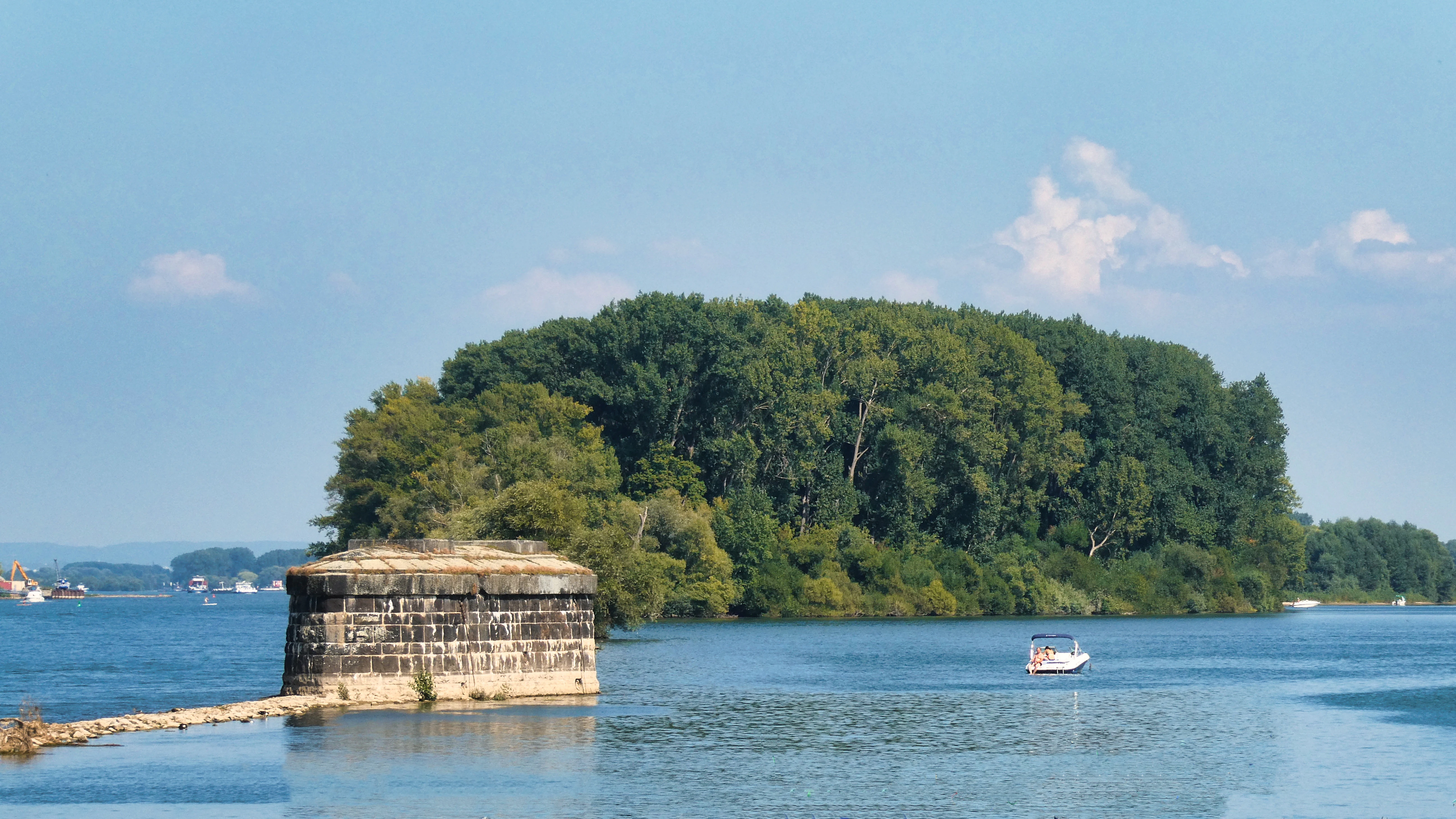 A view of a lush, tree-covered river island in the Rhine, with a prominent stone bridge pier in the foreground and a small boat on the water.