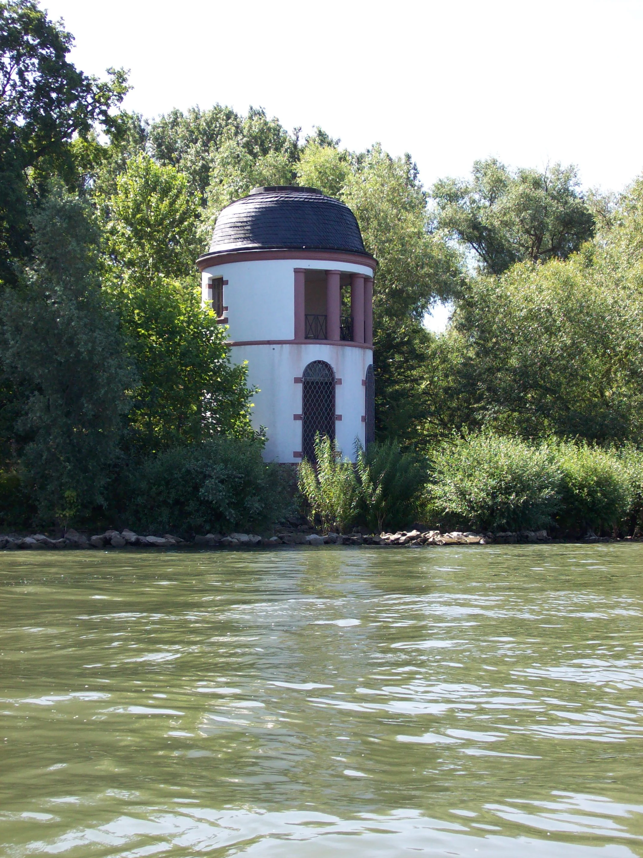 A white, circular stone observation tower with a dark dome roof sits along the tree-lined riverbank of the Rhine.