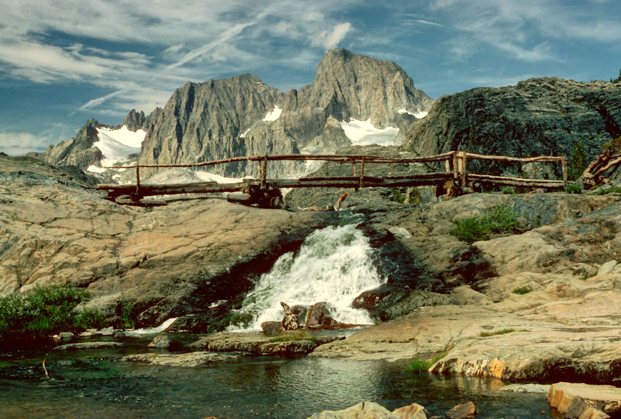 A rustic log footbridge crosses a small waterfall cascading over rocky terrain, with prominent snow-capped jagged mountain peaks in the distance.