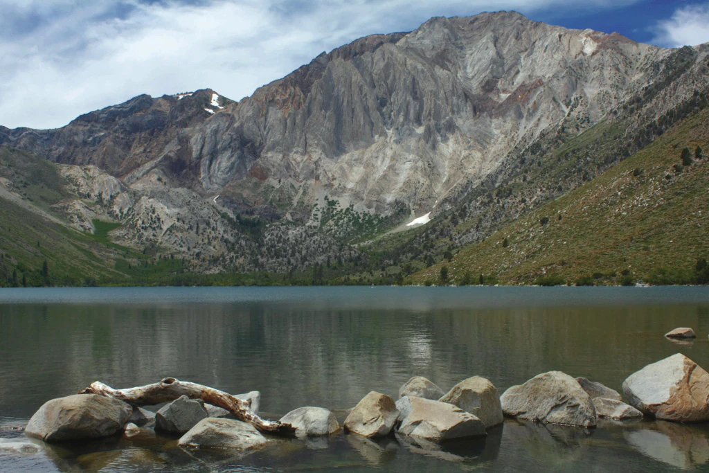 A wide view of a large, steep, rocky mountain reflecting in a clear lake, with scattered stones and a fallen branch along the foreground shoreline.