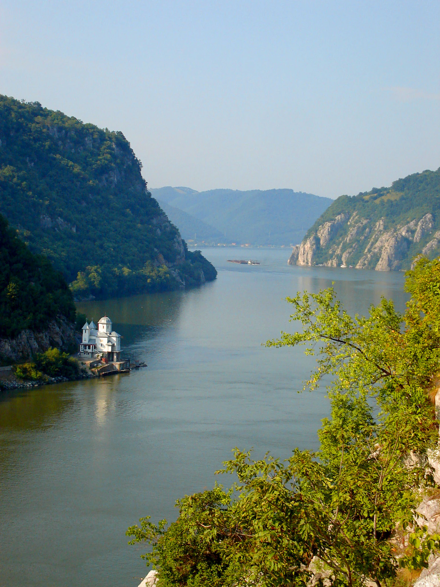 Wide view of the Danube River between steep green-clad cliffs, featuring a small white building on the riverbank and a boat in the distance
