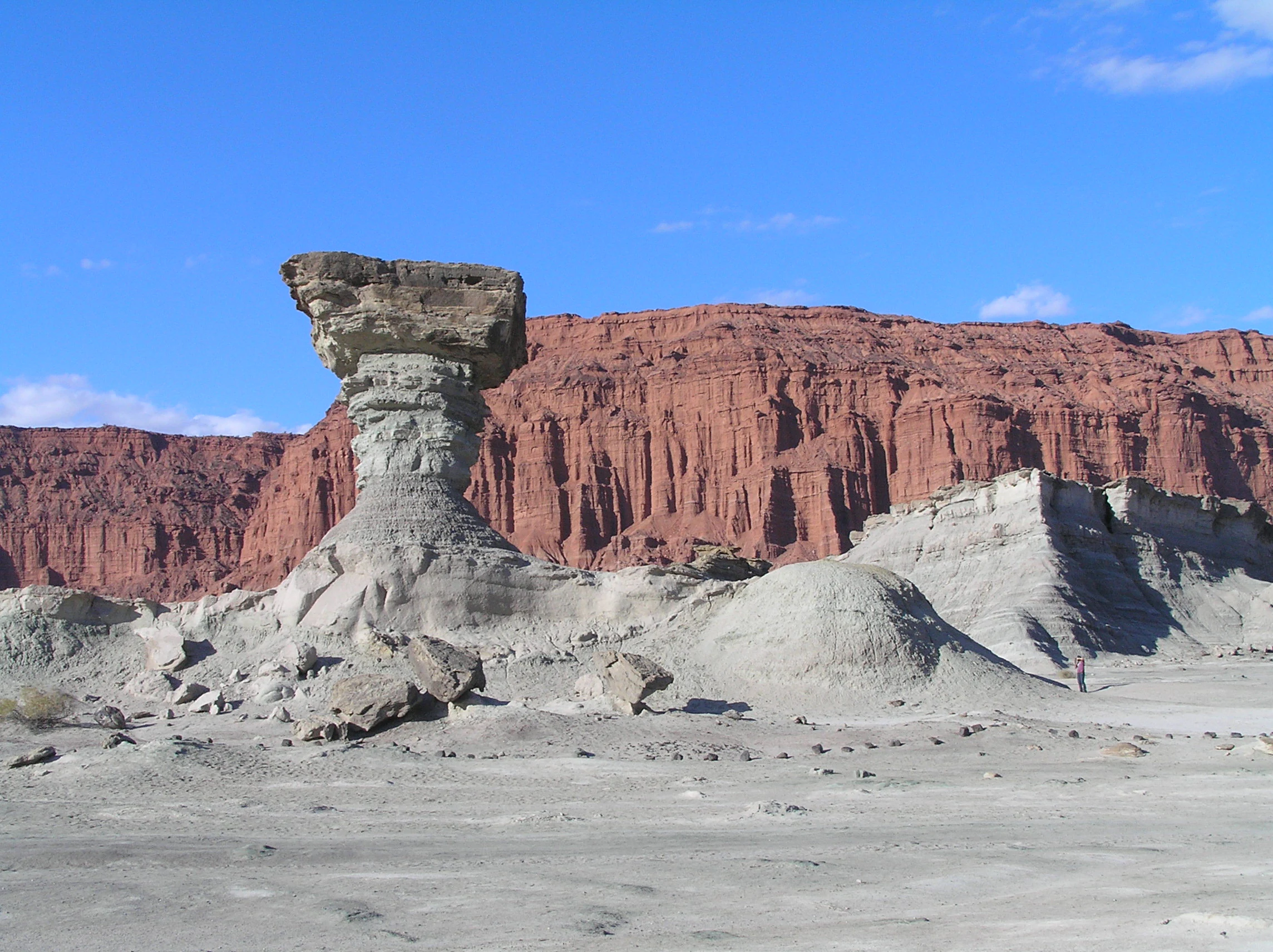 A large, weathered, mushroom-shaped rock formation stands in a desolate, pale grey desert landscape, with a towering, layered red canyon cliff in the background under a clear blue sky.