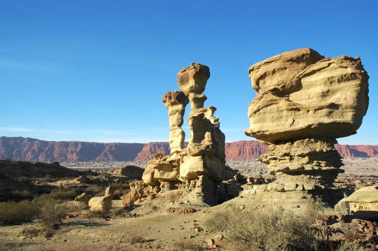 A desert landscape at Ischigualasto Provincial Park featuring towering, uniquely eroded sandstone rock formations under a clear blue sky, with a long red mountain ridge in the background.