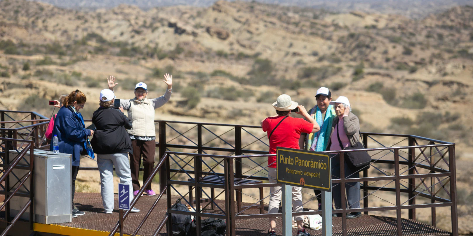 A group of tourists standing on a metal observation deck with a sign reading Punto Panoramico, looking out over a dry, hilly desert landscape.