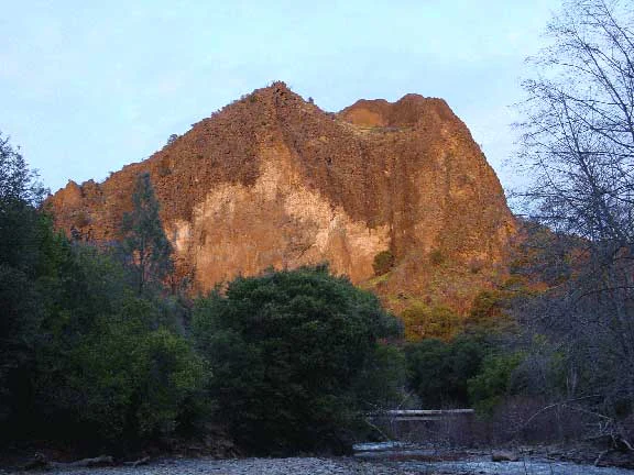 A large, craggy, reddish-brown rock formation rising above a forest canopy and a riverbed in the Ishi Wilderness.