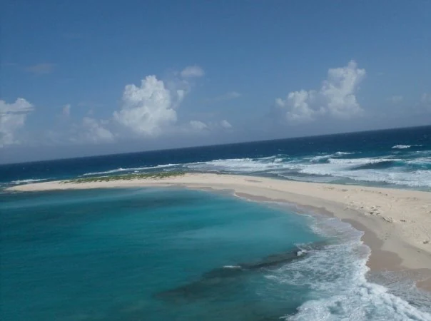 Una vista aérea que muestra una isla arenosa estrecha en forma de media luna rodeada de aguas oceánicas turquesas y azul profundo.