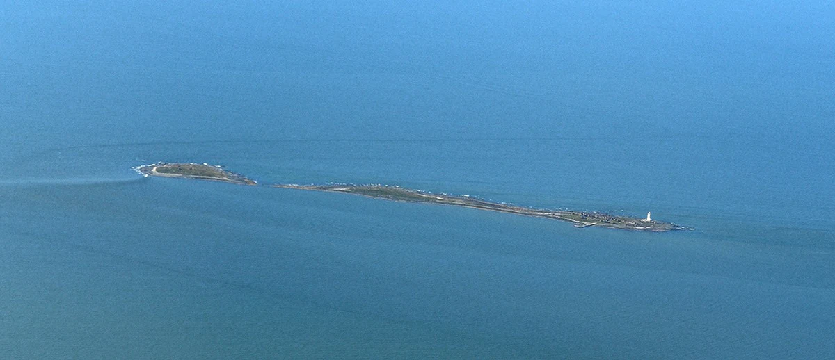 An aerial shot of the long, narrow Isla de Flores surrounded by the deep blue waters of the Rio de la Plata, with a small white lighthouse visible at one end of the island.