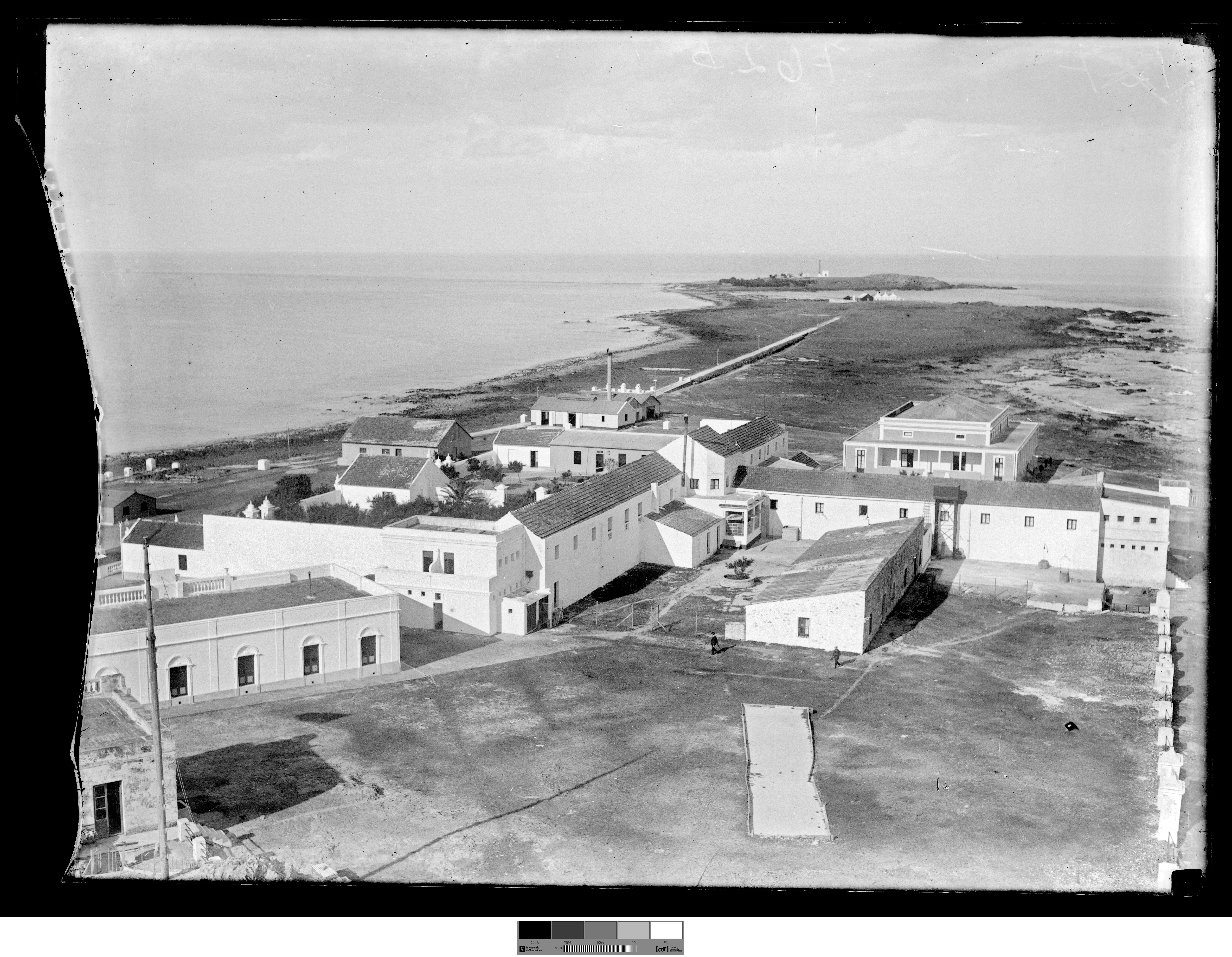 An archival black and white photograph showing a complex of low, white stone buildings situated on a flat coastal landscape, with a path leading toward the horizon.