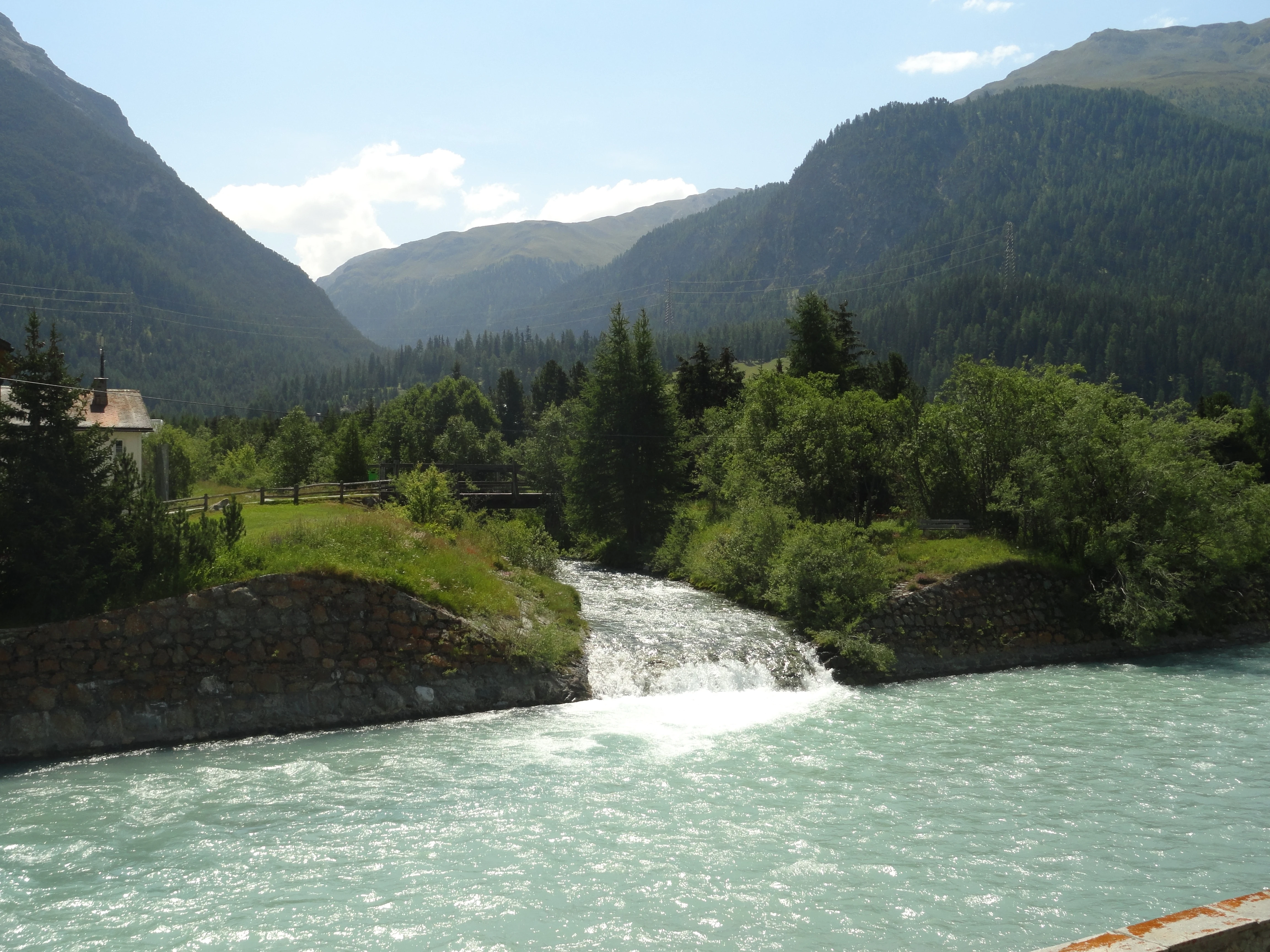 Una confluencia de ríos de montaña que muestra un arroyo más pequeño y de caudal rápido que desemboca en un río glaciar más grande y de color azul claro, con bosques alpinos y laderas de valle circundantes.