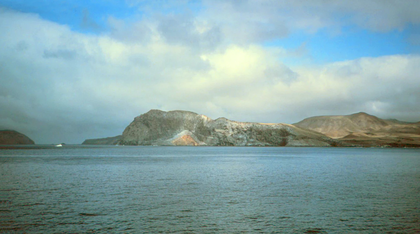 A wide coastal view of the rocky, barren Isla Guadalupe island with calm waters in the foreground and a cloudy blue sky above.