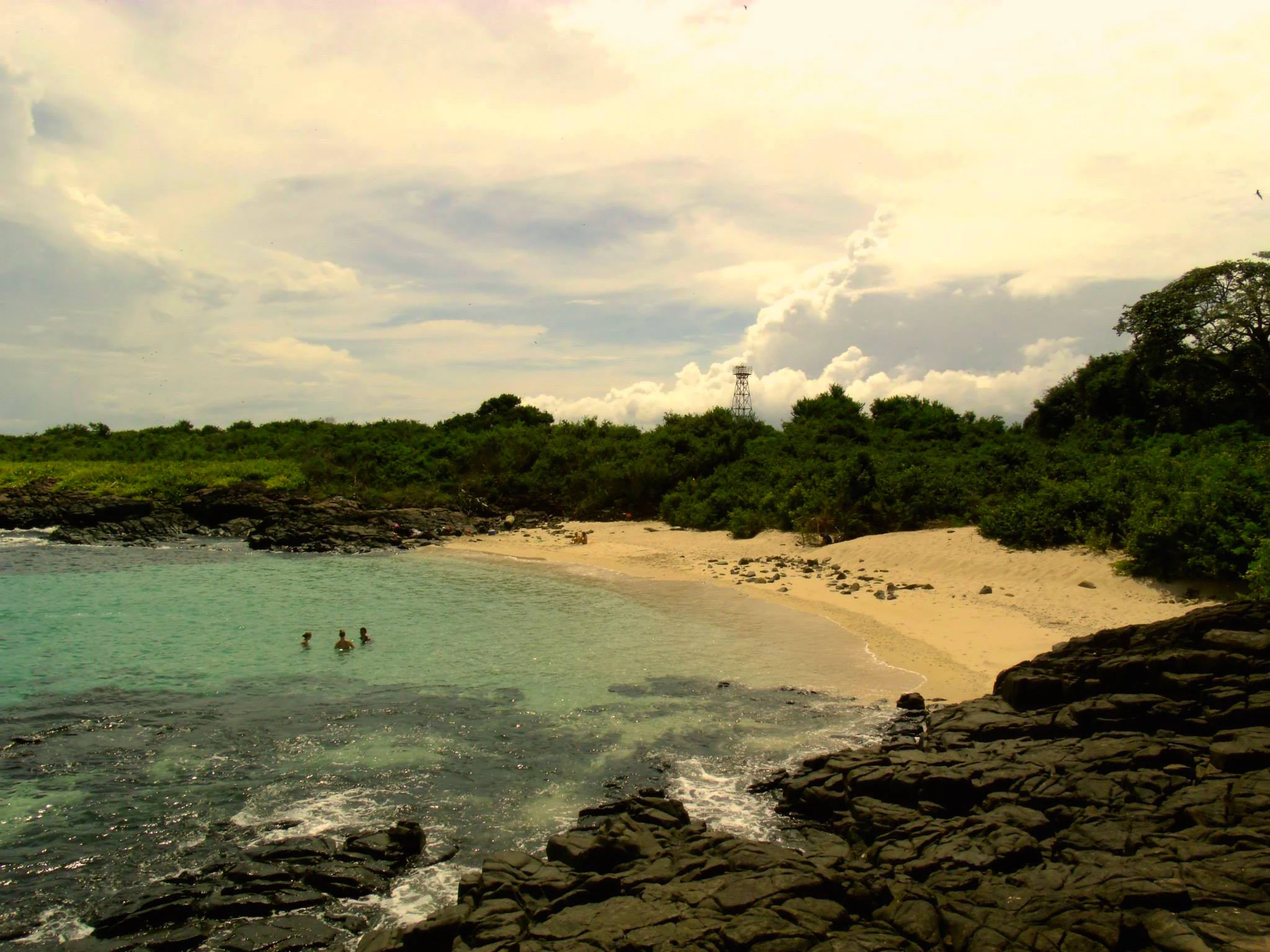 A sandy beach with dark volcanic rocks in the foreground and turquoise water with people swimming, backed by a lush green forest under a cloudy sky.
