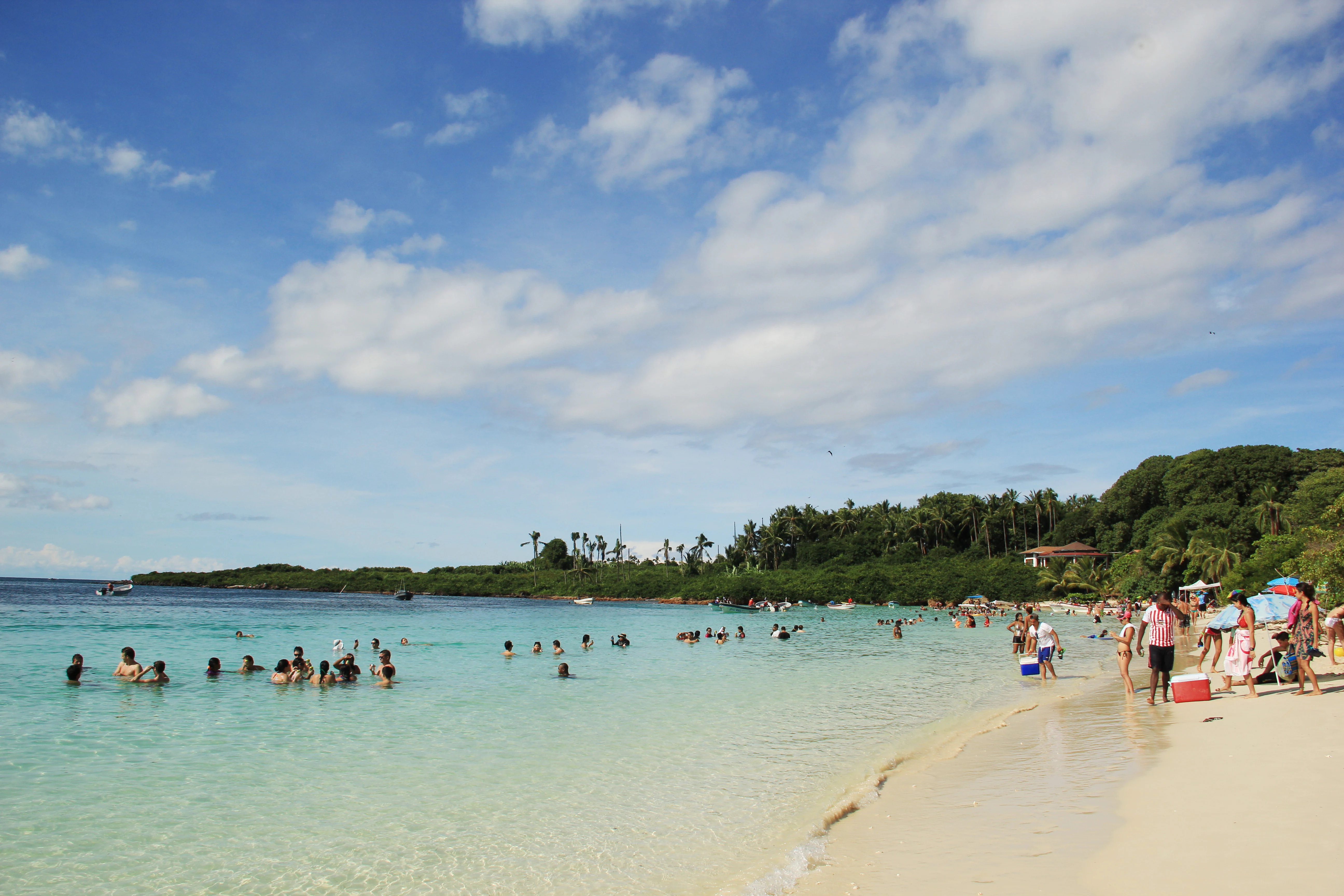 A sunny beach scene on Isla Iguana with tourists wading and swimming in turquoise water, framed by lush green trees and a white sandy shore under a bright blue sky with scattered clouds.