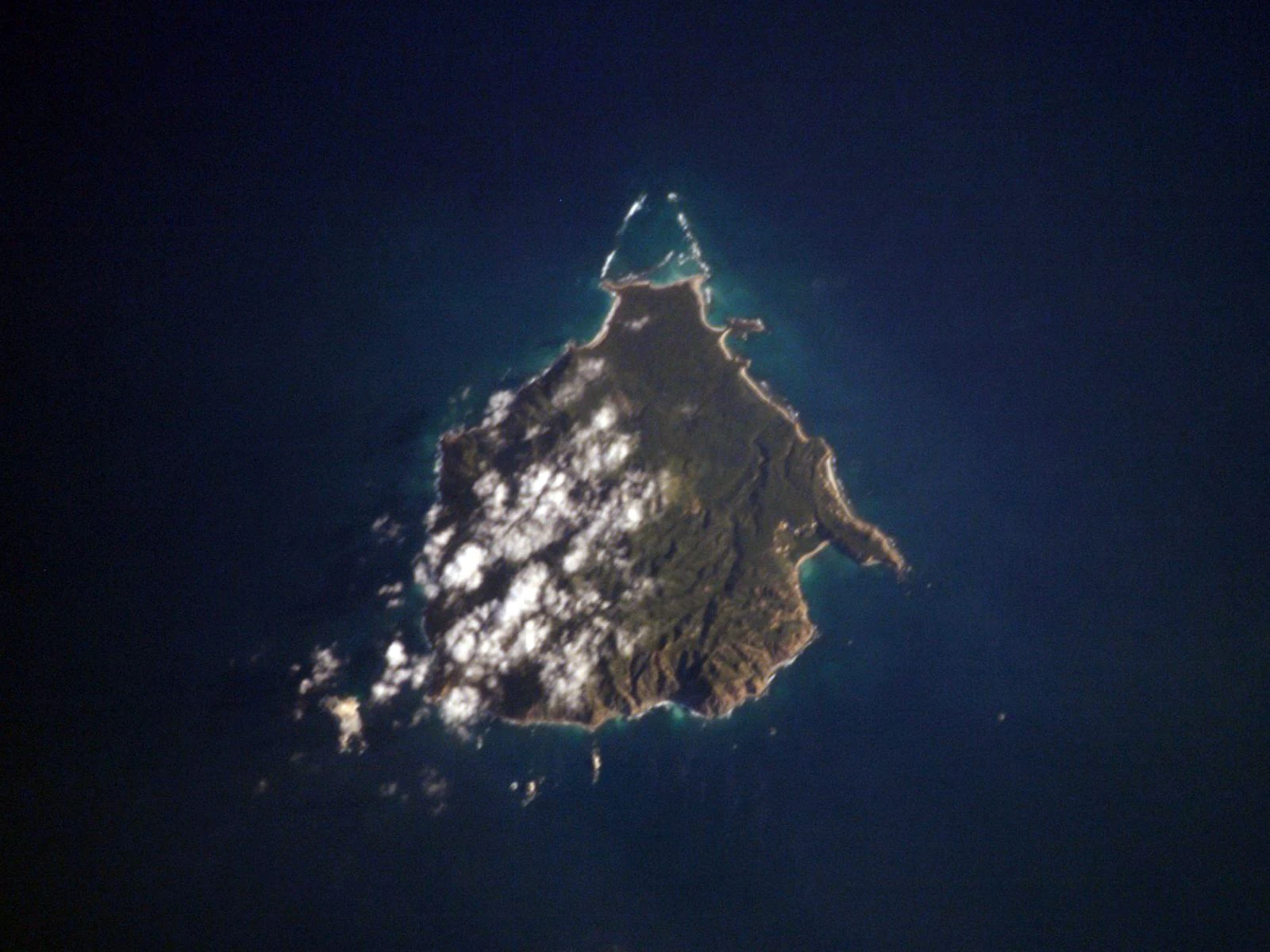 Una fotografía satelital cenital de la isla María Cleofas rodeada de aguas oceánicas azul profundo, con nubes dispersas proyectando sombras sobre la superficie de la isla.