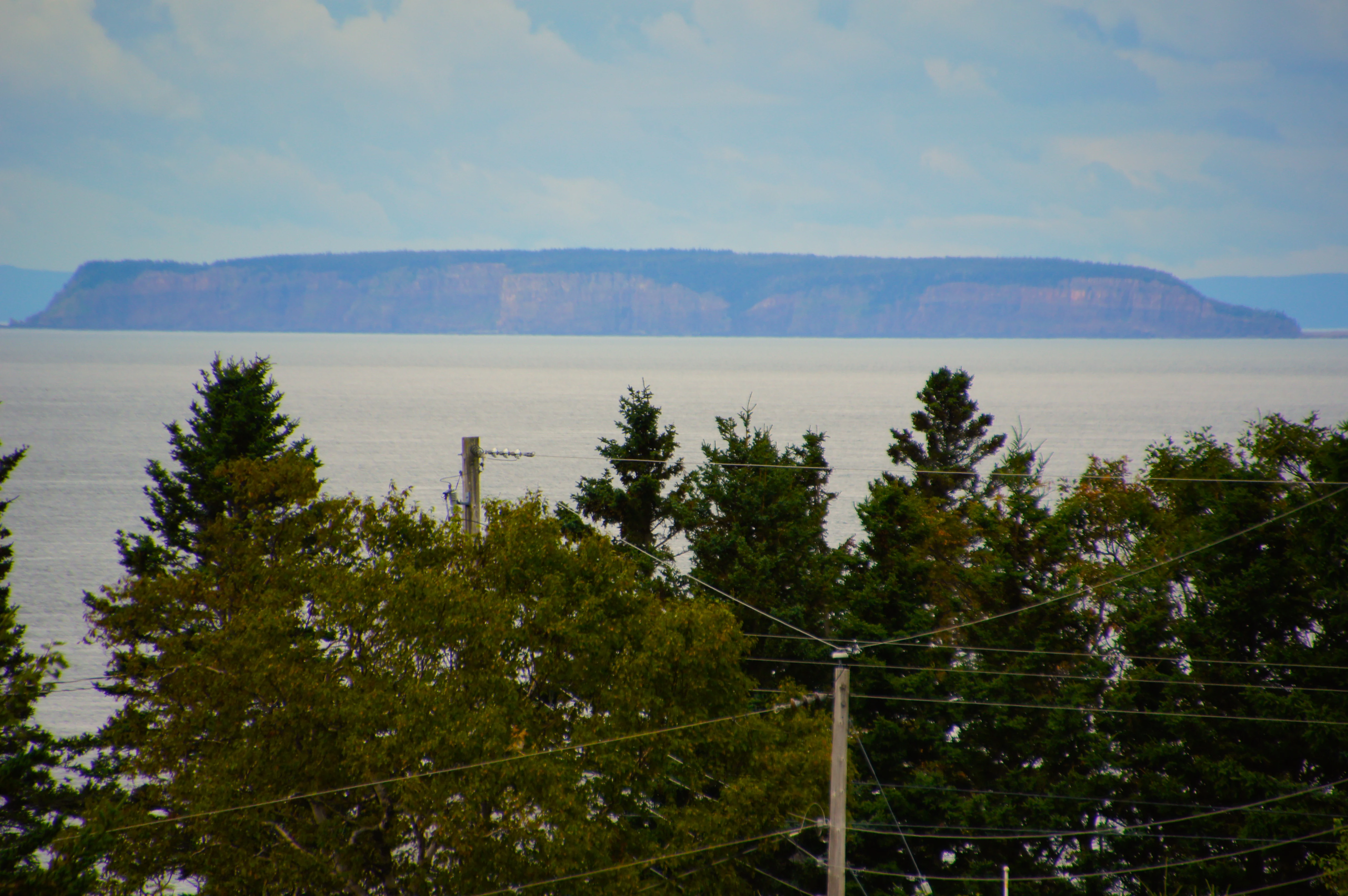A horizontal, long-distance view of the flat-topped Isle Haute island rising above the water of the Bay of Fundy, framed by foreground trees and power lines.