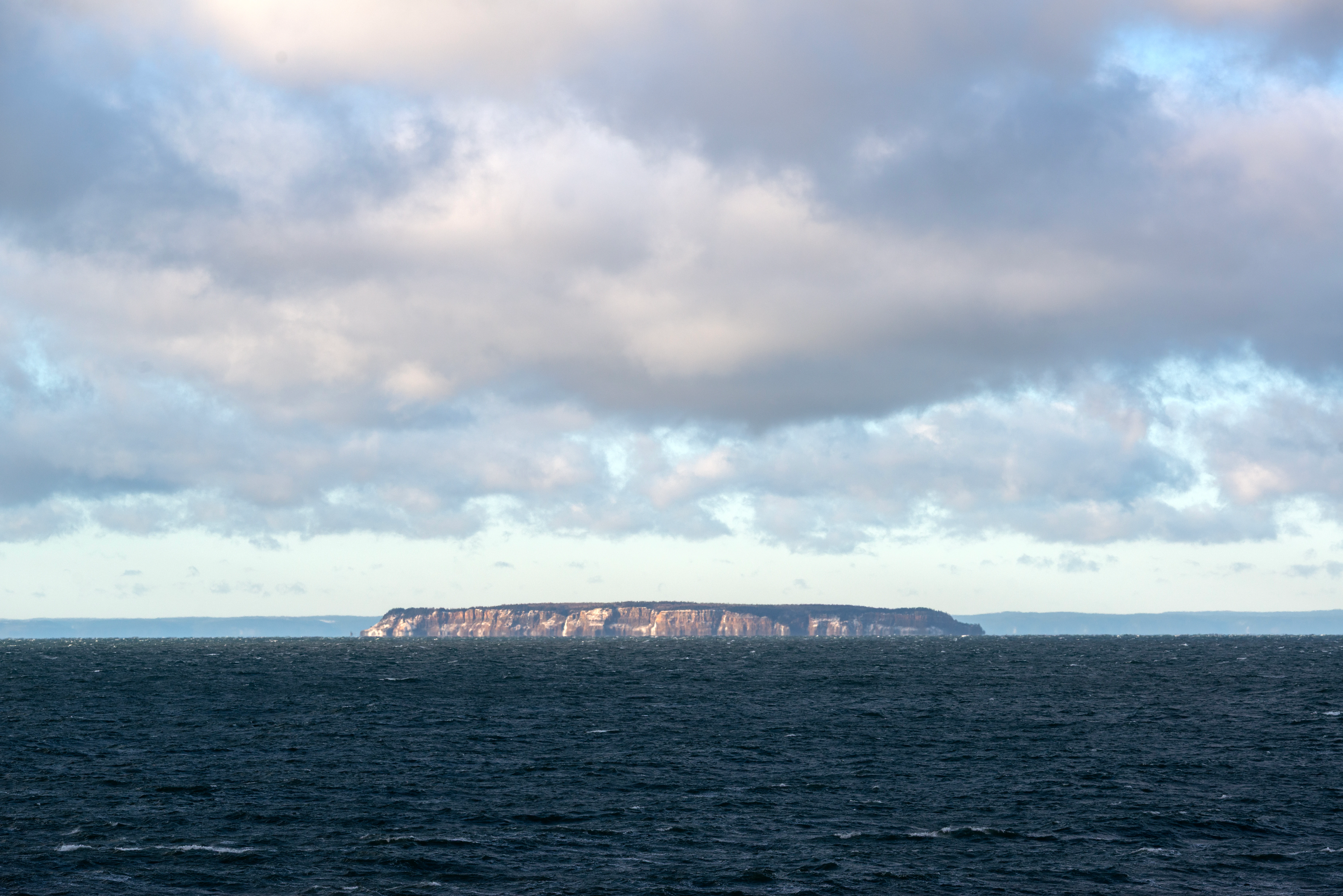 A distant, low-angle shot of a rocky island with flat-topped cliffs rising from a dark, choppy ocean under a cloudy sky.