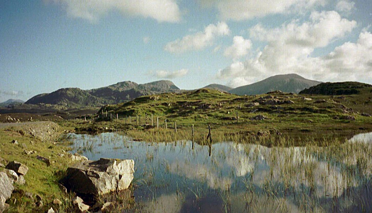 A landscape view of a loch in the foreground with grassy, rocky terrain and rolling mountains in the distance under a partly cloudy sky.