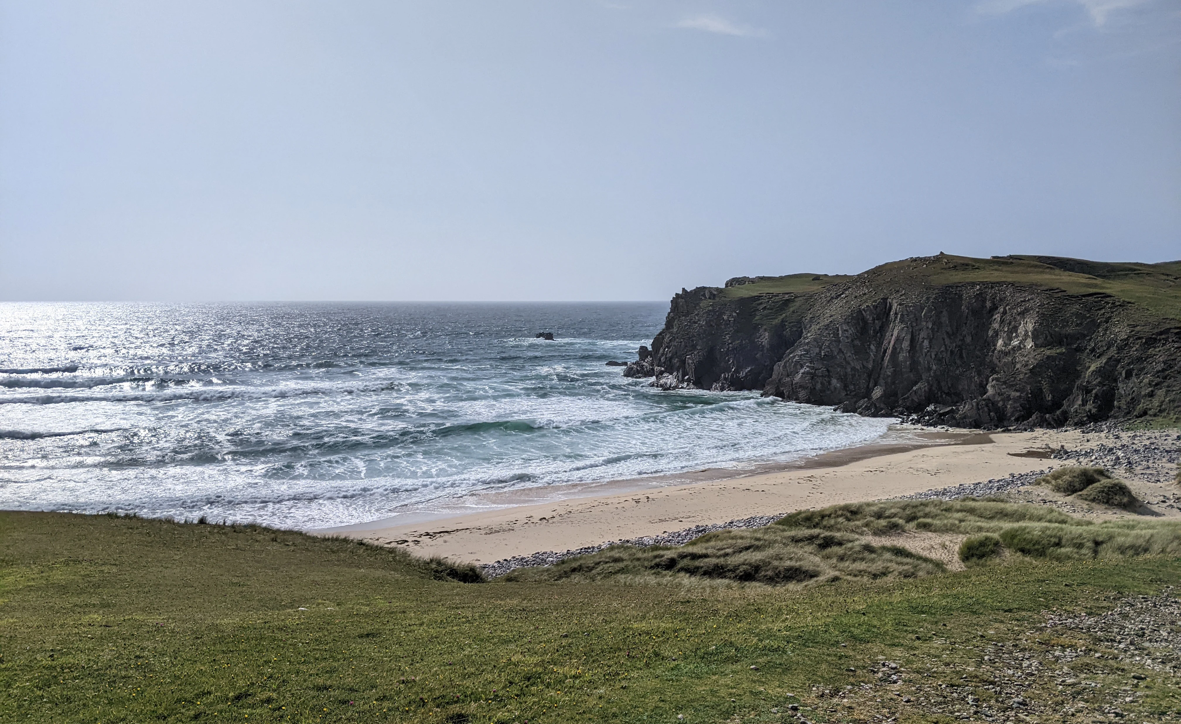 A wide angle view of a sandy beach bordered by steep dark cliffs and grassy dunes on a bright day.