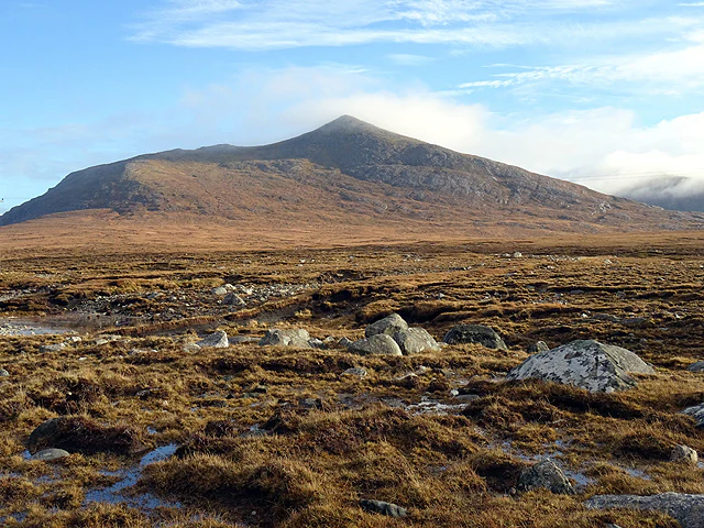 A wide shot of a vast, brown, boggy landscape covered in moss and scattered stones, leading to a prominent, smooth-sloped mountain under a clear blue sky.