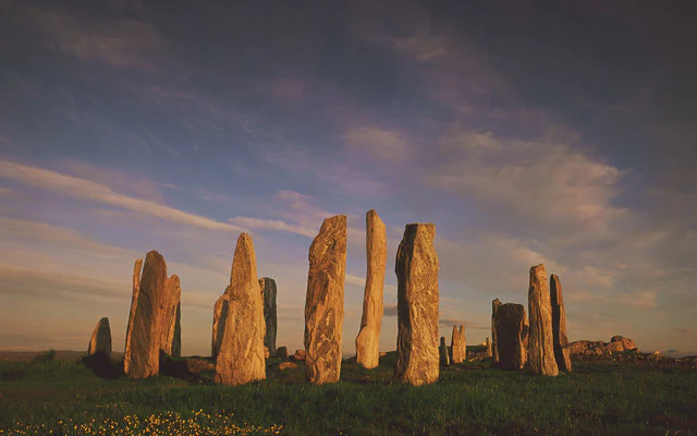 A group of tall, weathered standing stones arranged in a circle on a grassy hillside, illuminated by the warm light of a late sunset.