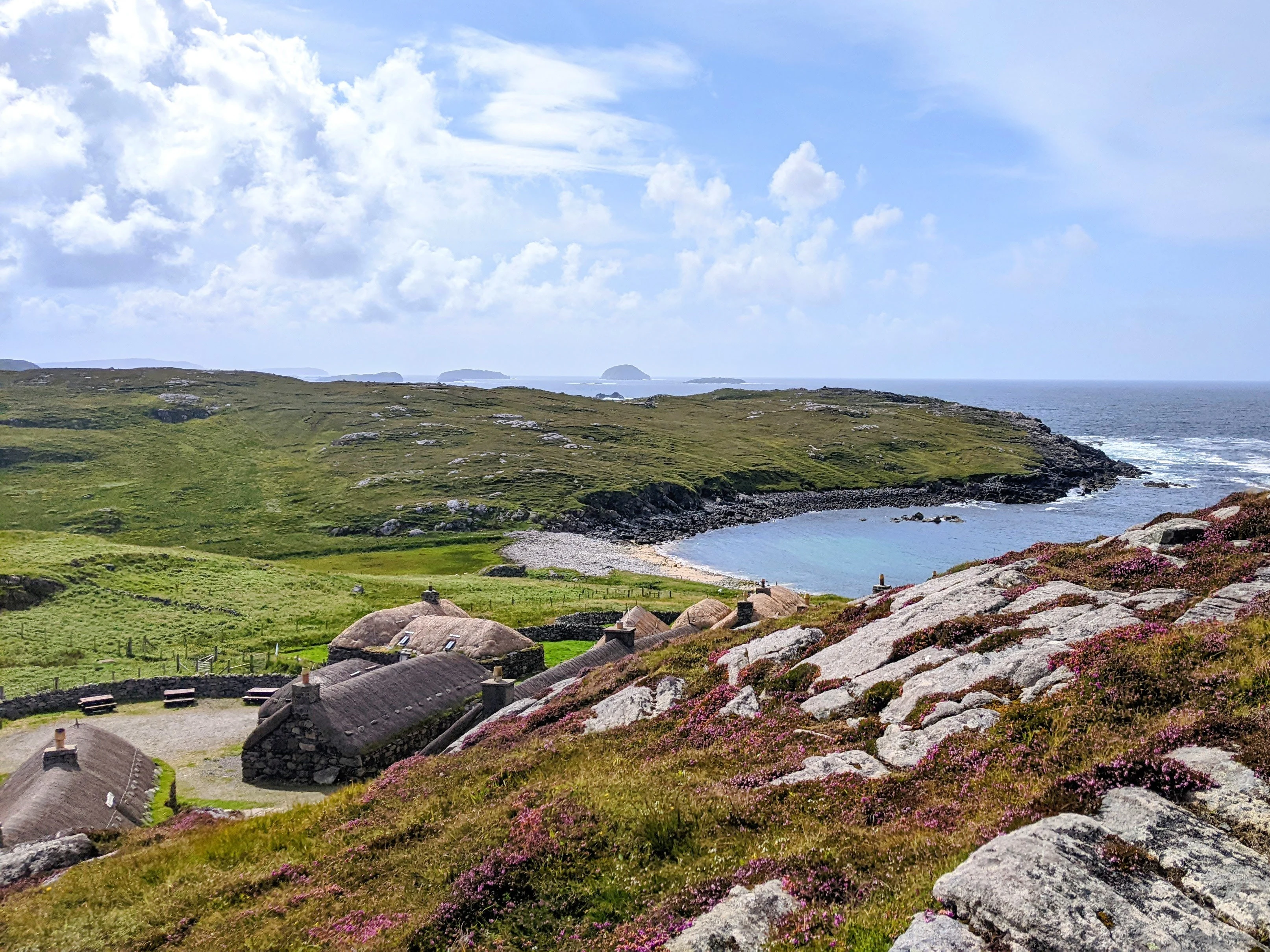 A high-angle view of traditional stone cottages with thatched roofs nestled in a green valley near a rocky coastline, with the ocean in the background under a partly cloudy sky.