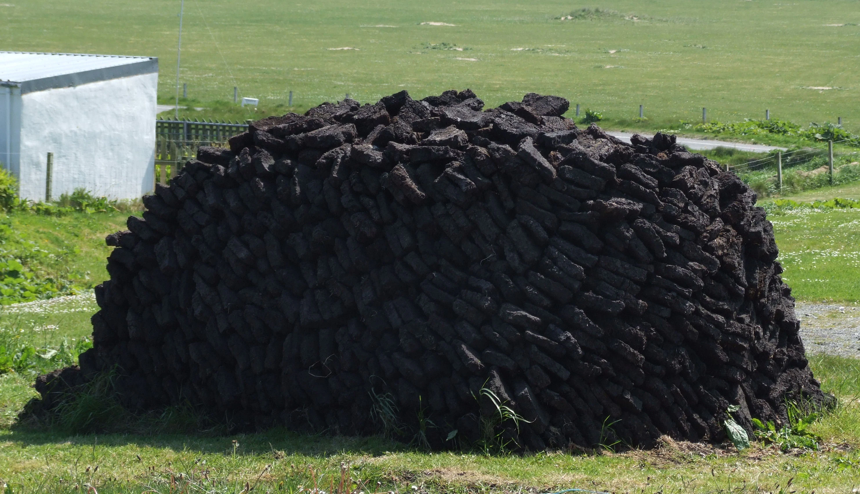 A large, rounded, neatly organized stack of cut, dark brown peat blocks resting on a green grassy field, with a glimpse of a white building structure to the left.